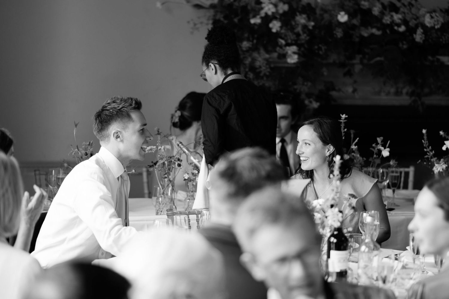 Black and white candid photo of wedding reception guests engaging in conversation at dinner tables.