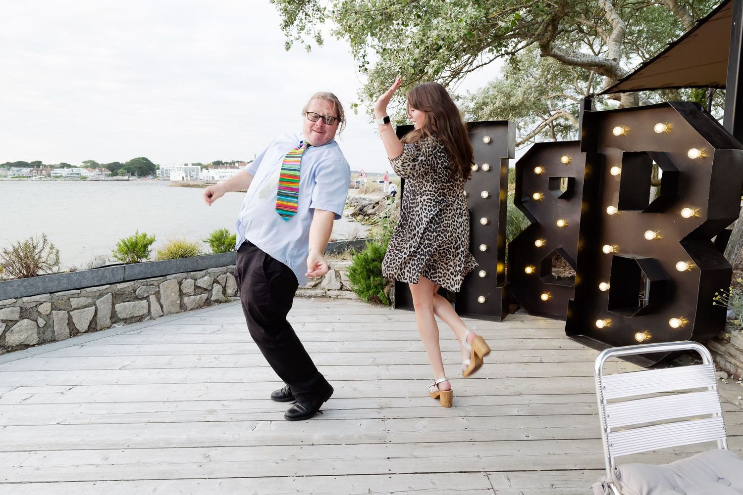 Playful dancing on wooden boardwalk by waterfront with 'R' marquee letter visible.