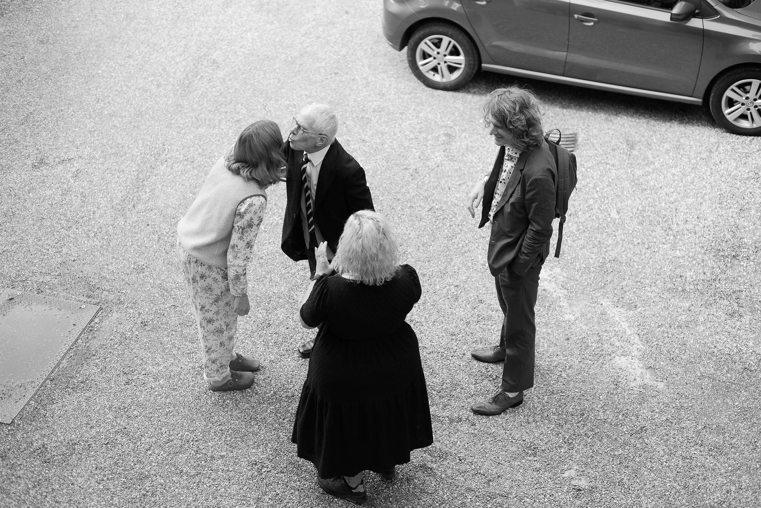 Wedding party gathering outdoors near parked cars on gravel surface.