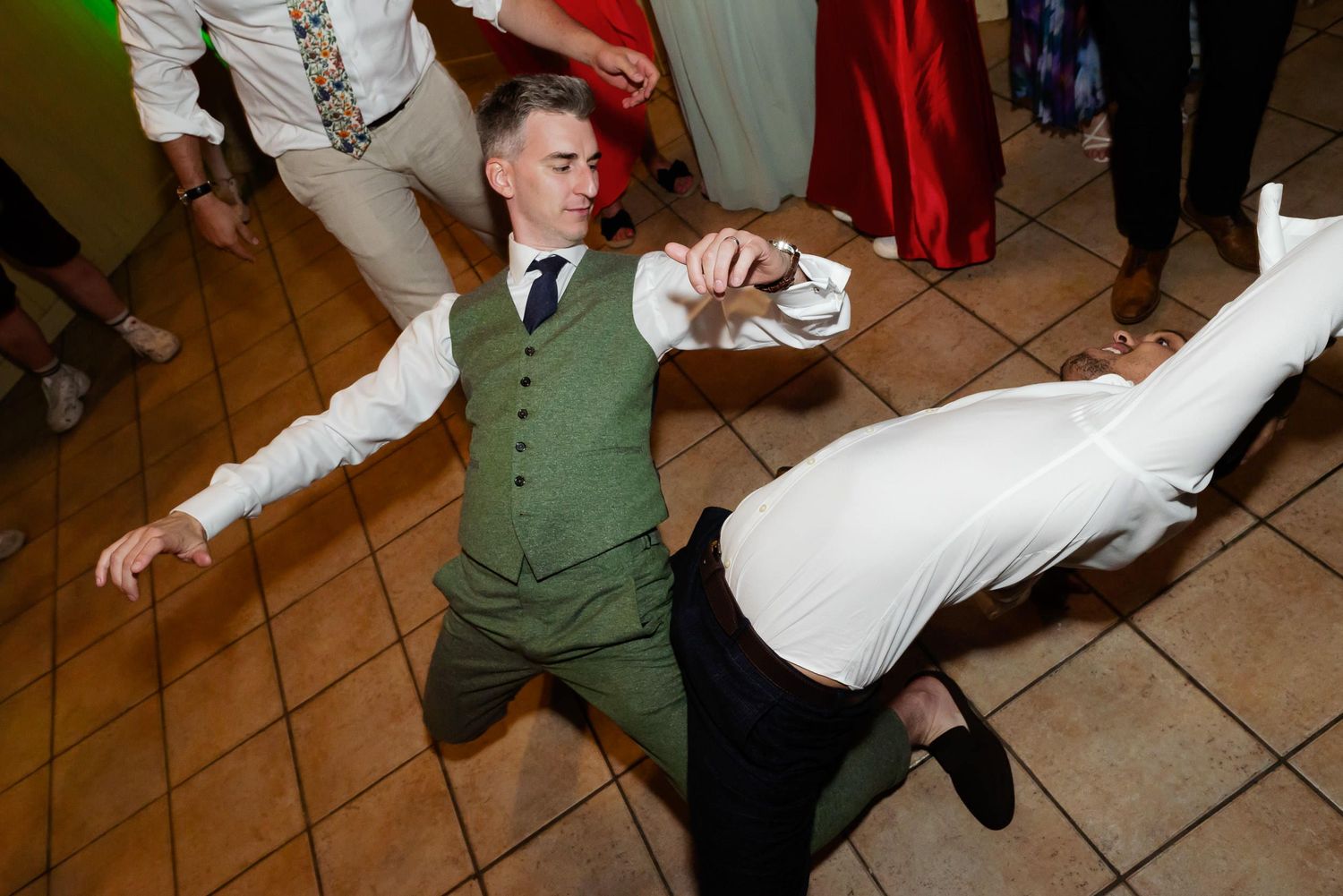Wedding guests perform dramatic dance moves at a reception on a tiled floor.