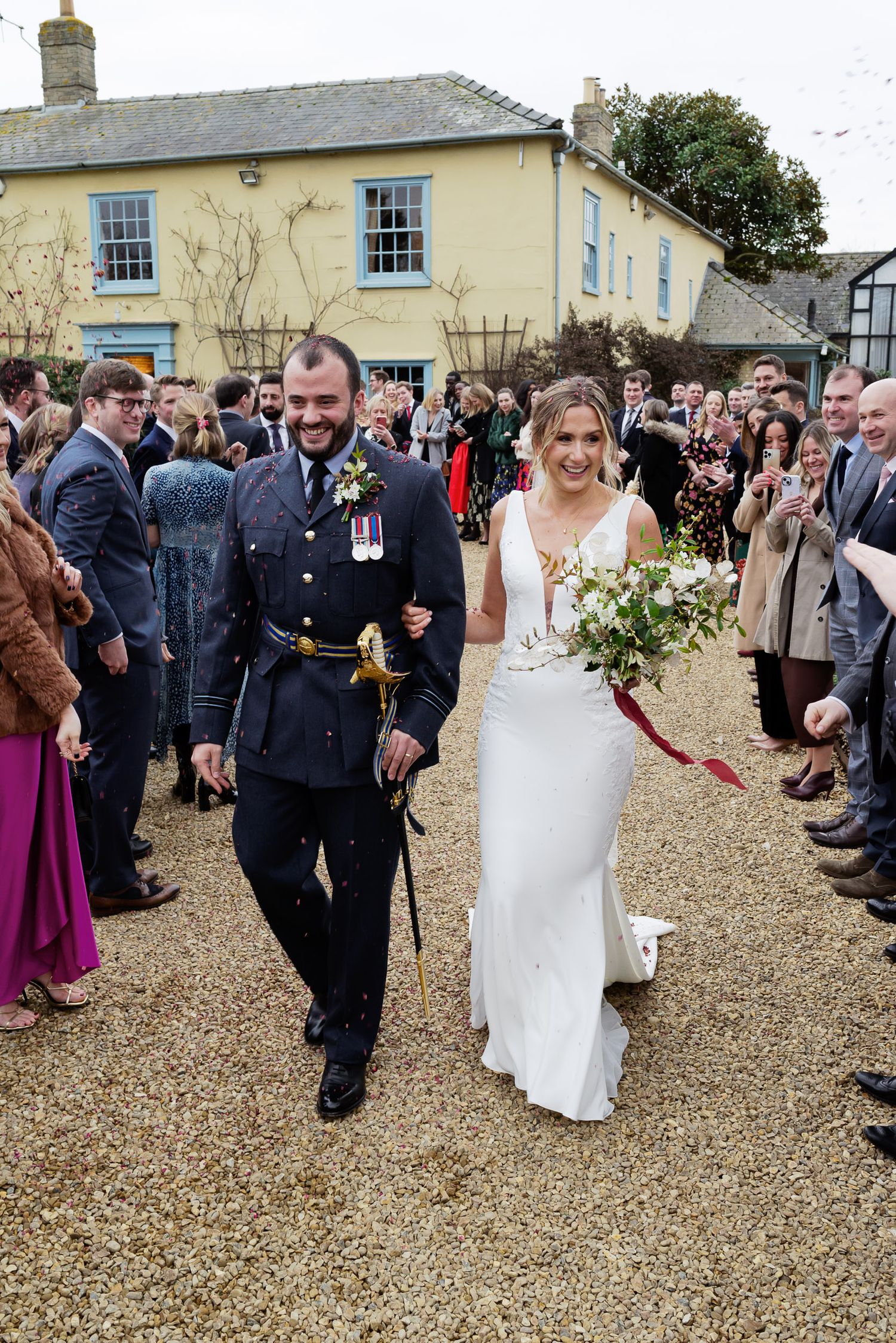 A military serviceman and bride walk through a crowd of wedding guests on a gravel path outside a yellow house.