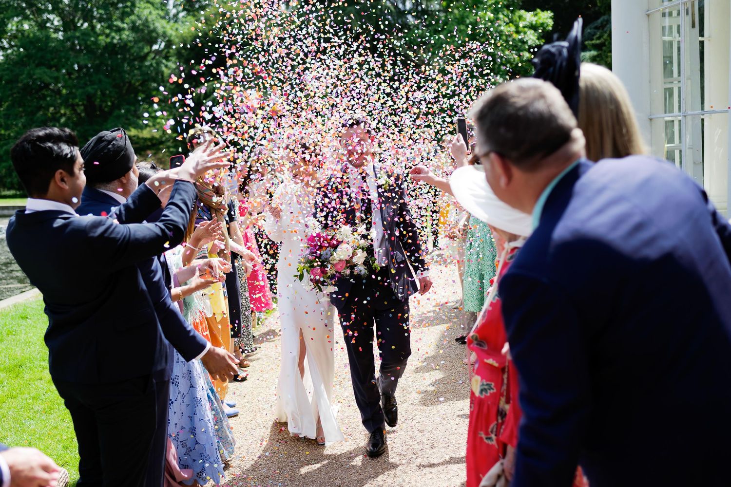 Wedding guests throw confetti over the newlyweds walking down garden path.