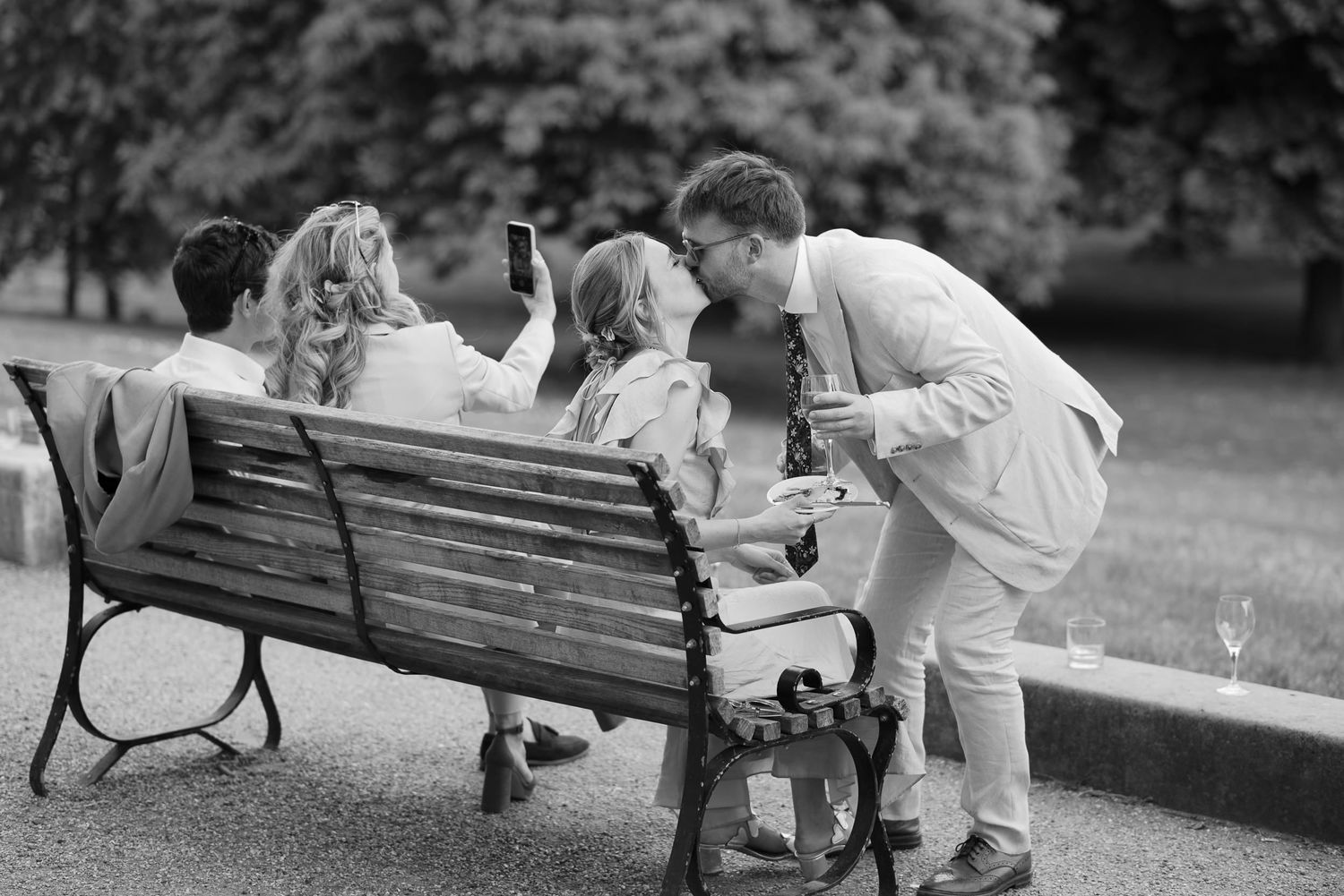A couple sits on a park bench in a playful moment as they lean away from each other while holding hands.