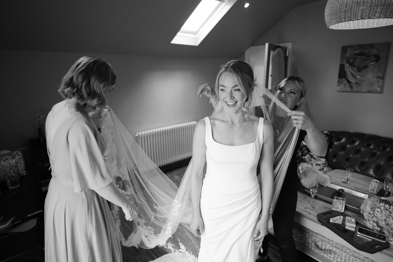Bride getting ready with help from others in a room filled with natural light and wedding preparations.