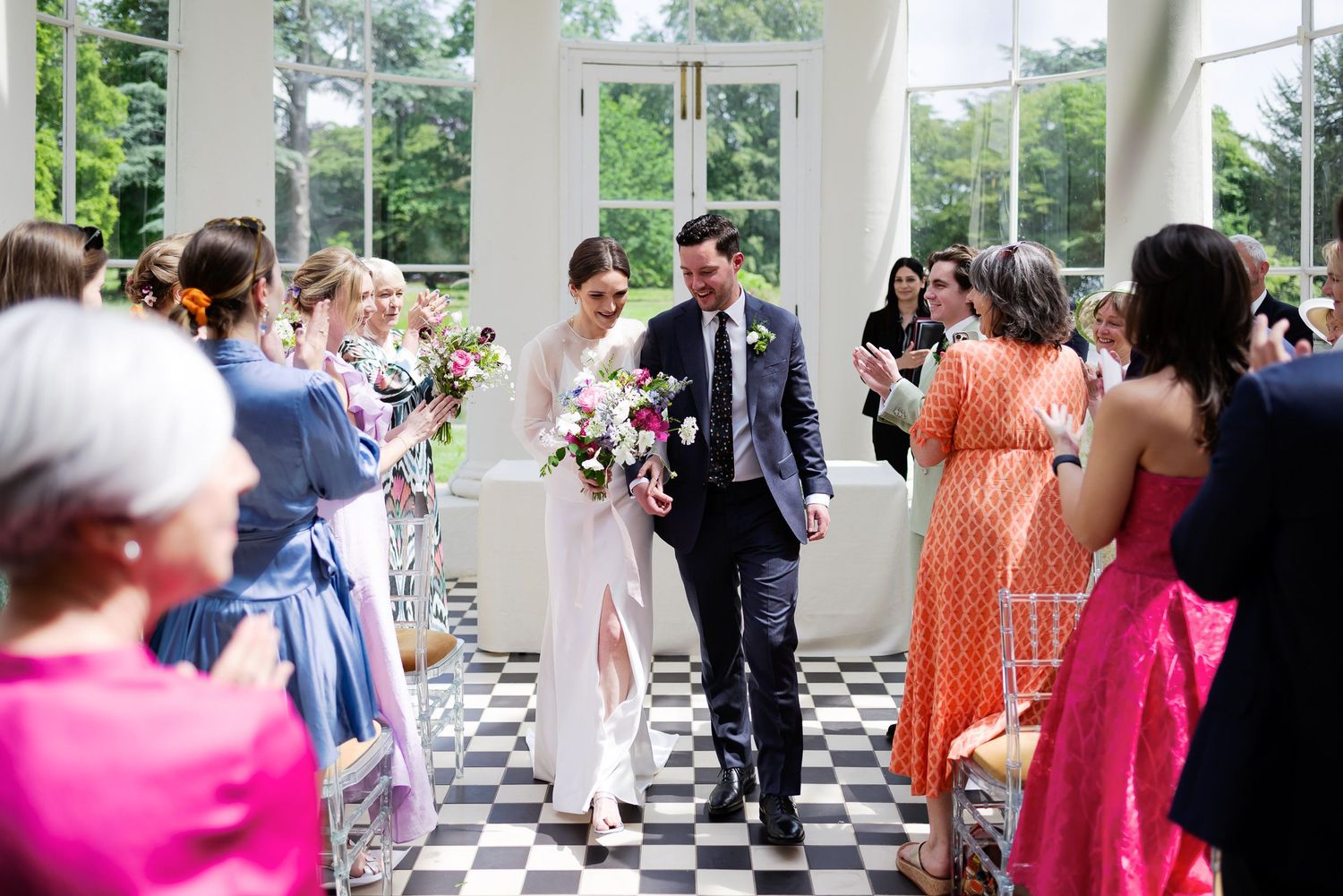 Wedding couple walking down aisle in bright conservatory with checkerboard floor and guests.