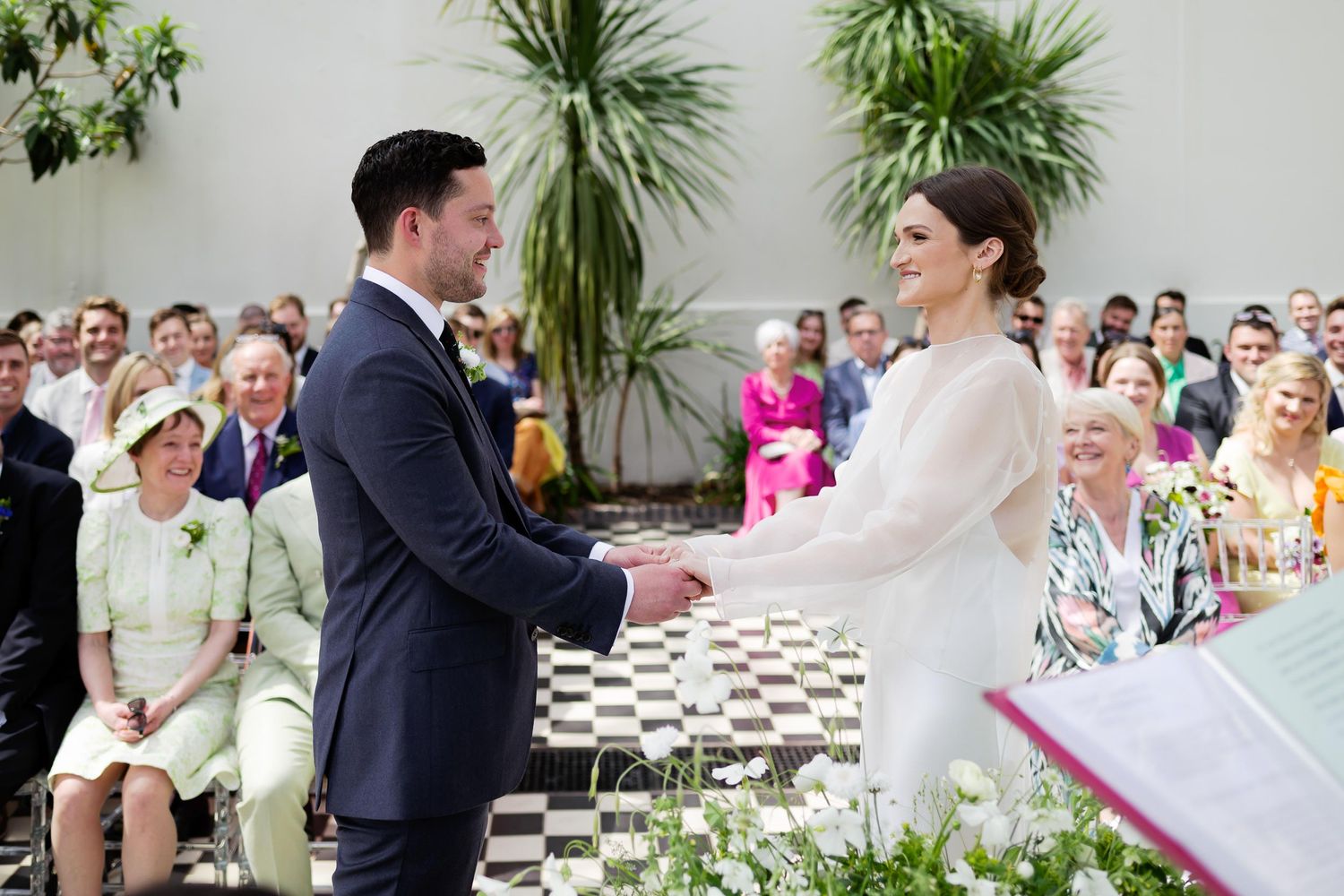 Wedding ceremony taking place indoors with tropical plants and guests watching the couple exchange vows.