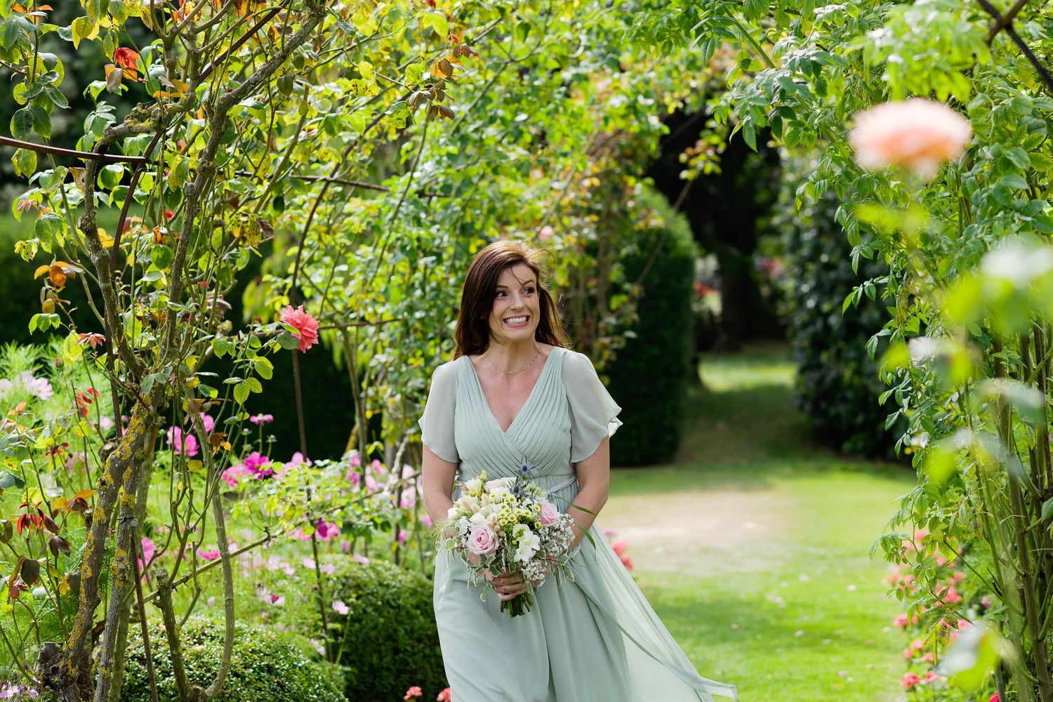 Woman in sage green dress walks through blooming garden holding wedding bouquet.