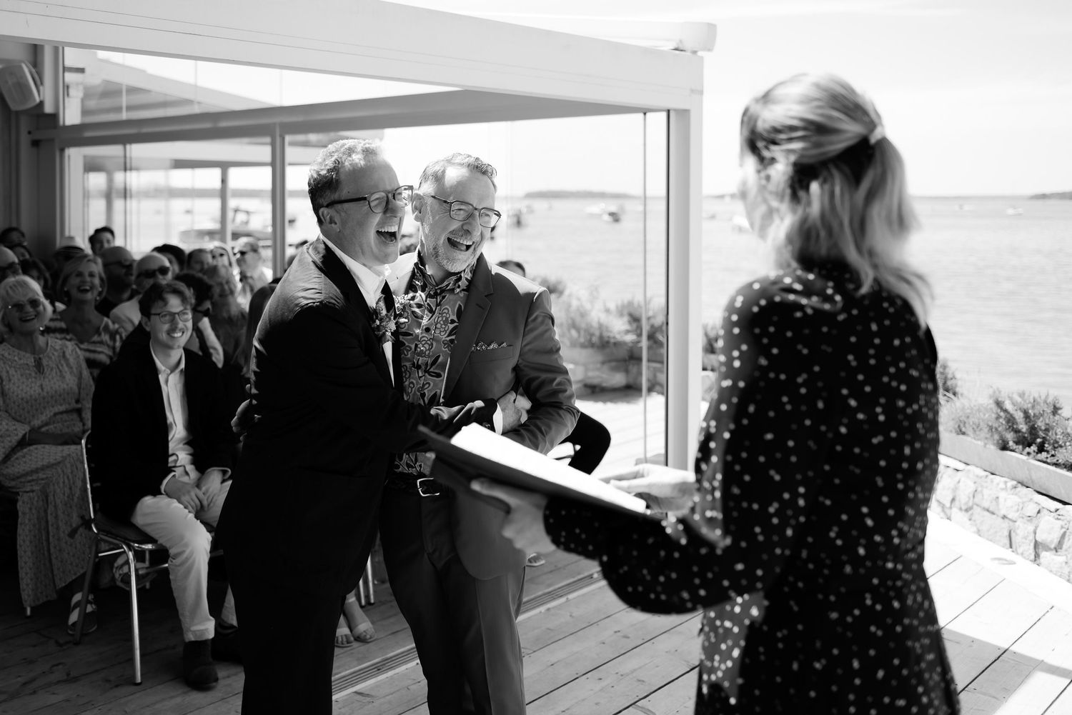 Black and white image of a ceremony in a bright seaside venue with large windows.