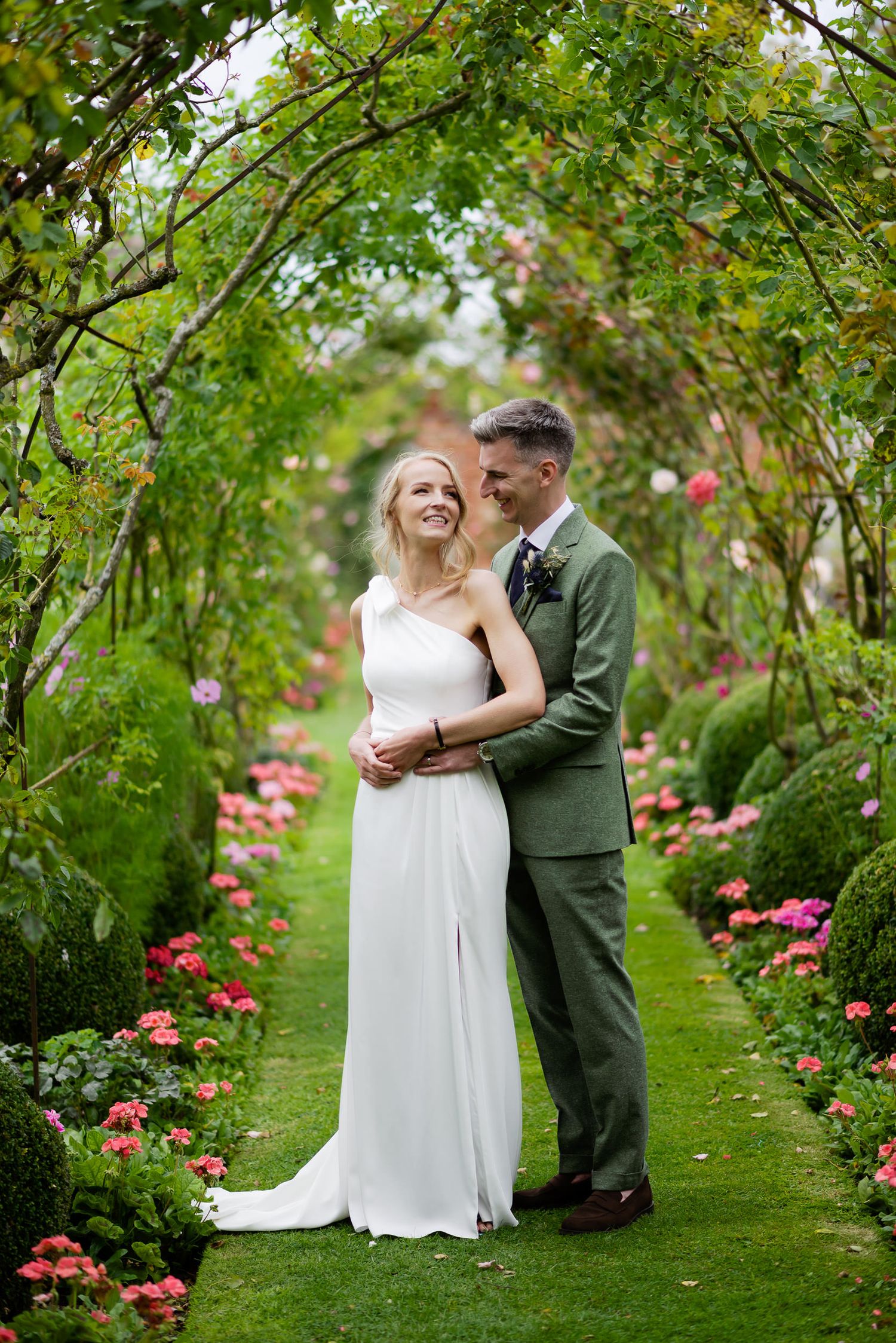 Couple embracing in a lush garden pathway lined with pink roses and climbing vines.