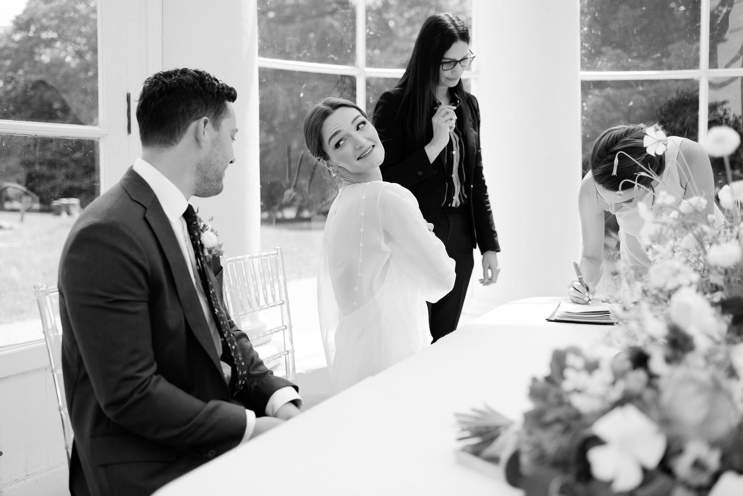 Black and white photo of a wedding couple signing documents at a table with floral decorations.