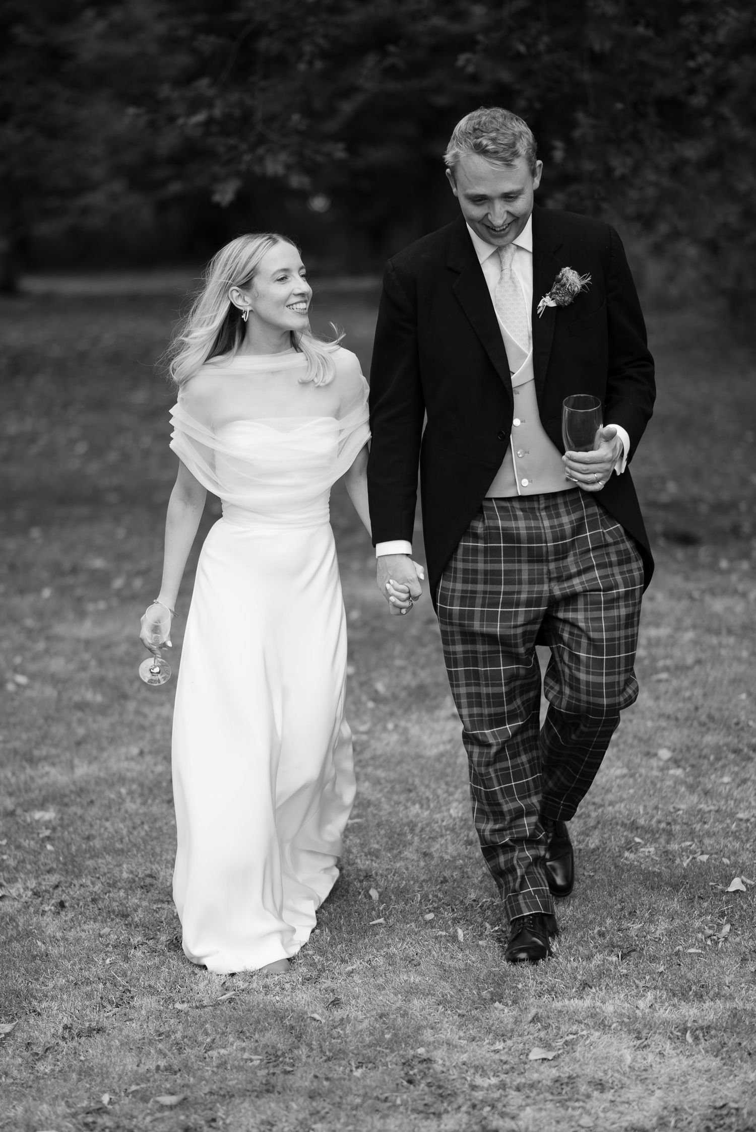 A black and white wedding photo of a couple walking outdoors, with the groom wearing plaid pants and bride in flowing dress.