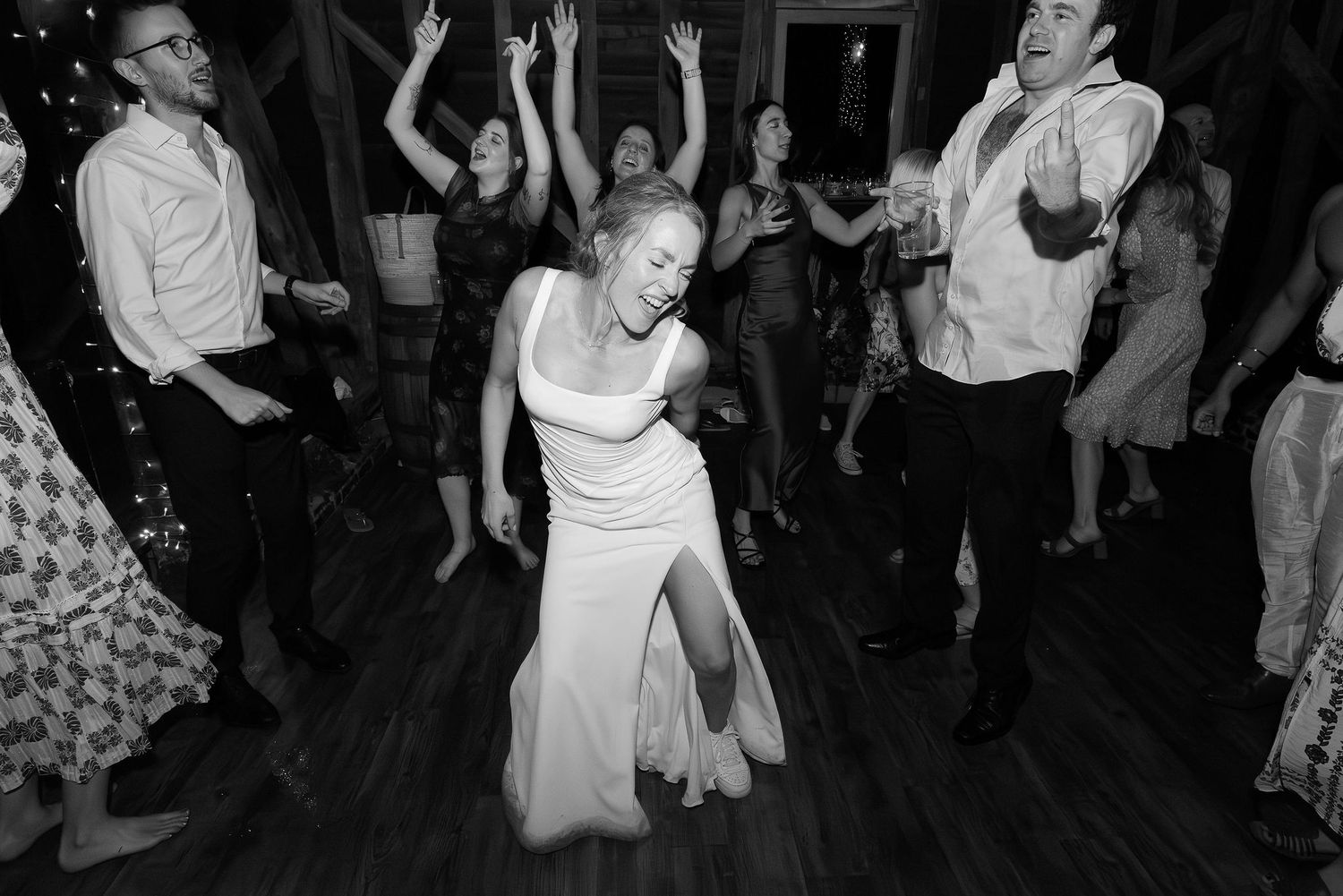 Black and white photo of wedding guests dancing with raised arms on a wooden floor.
