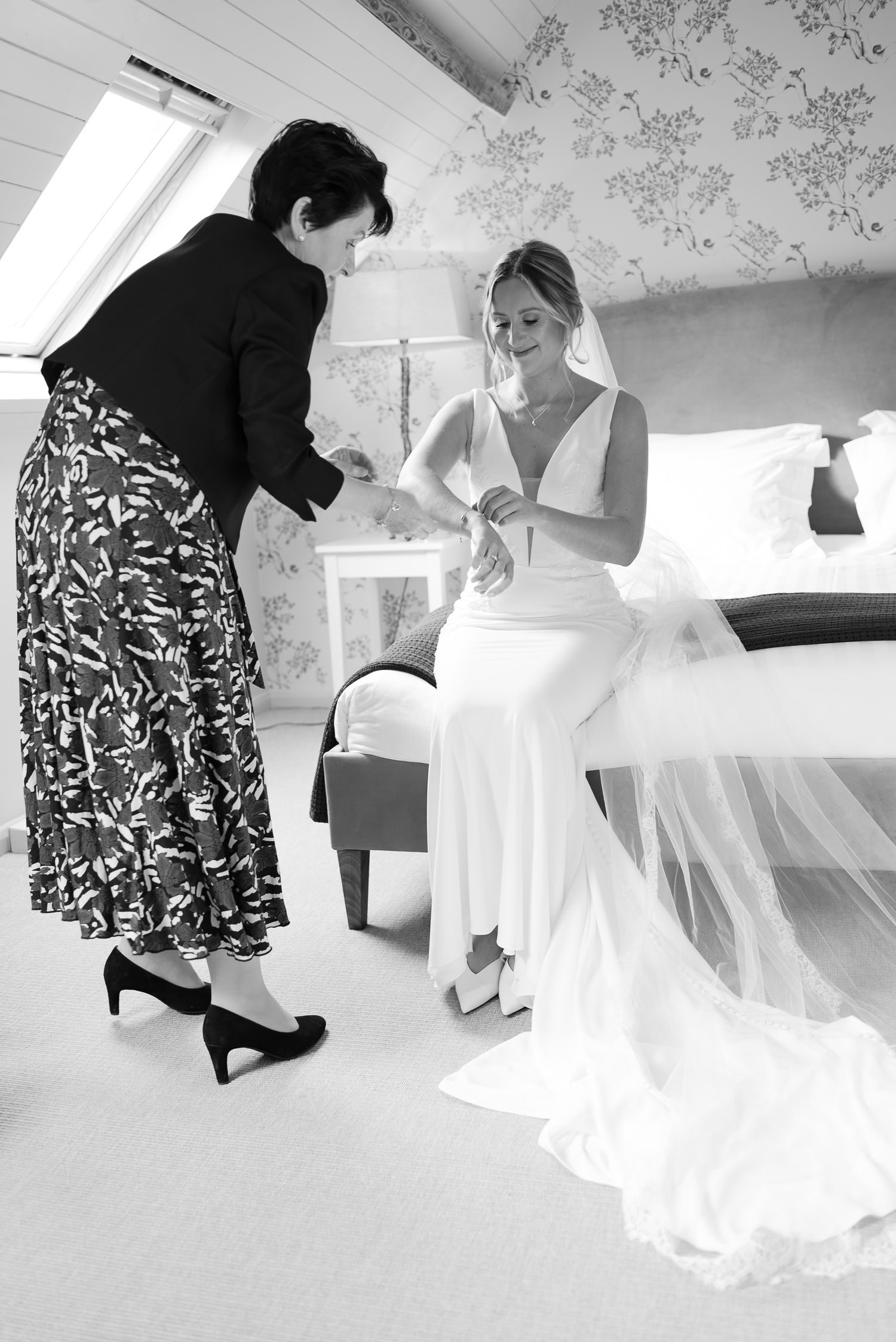 A series of black and white photos showing a bride getting ready with assistance in a room with floral wallpaper.