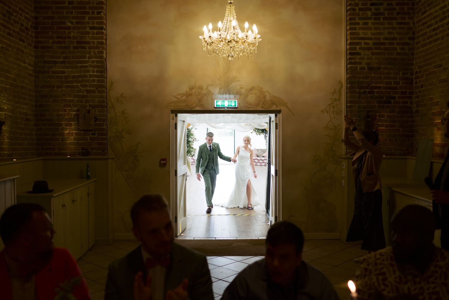A dimly lit elegant hallway with crystal chandeliers and brick walls showing silhouettes of people walking.