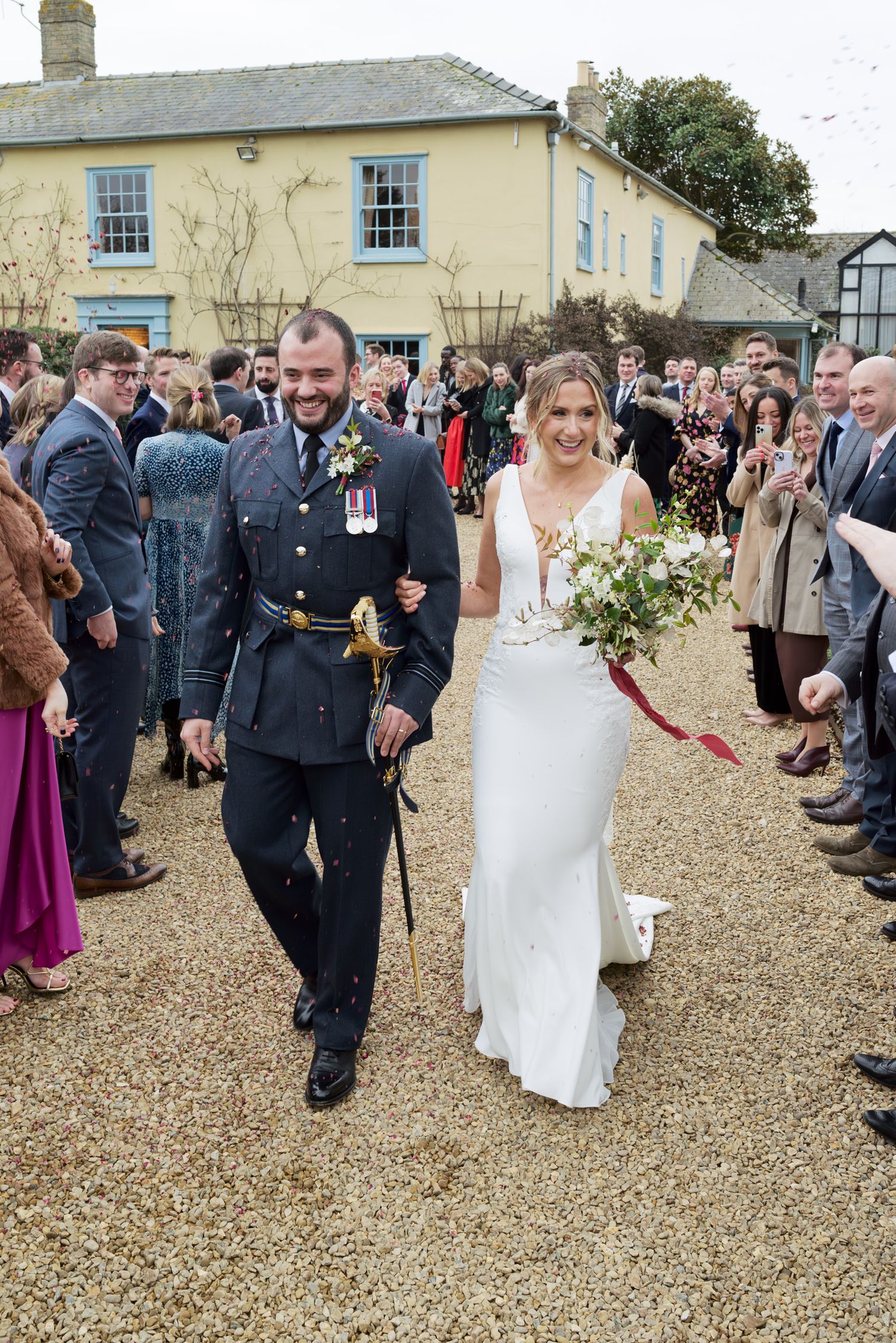 Wedding guests line up outside on a gravel path to toss confetti at a couple during their celebration.