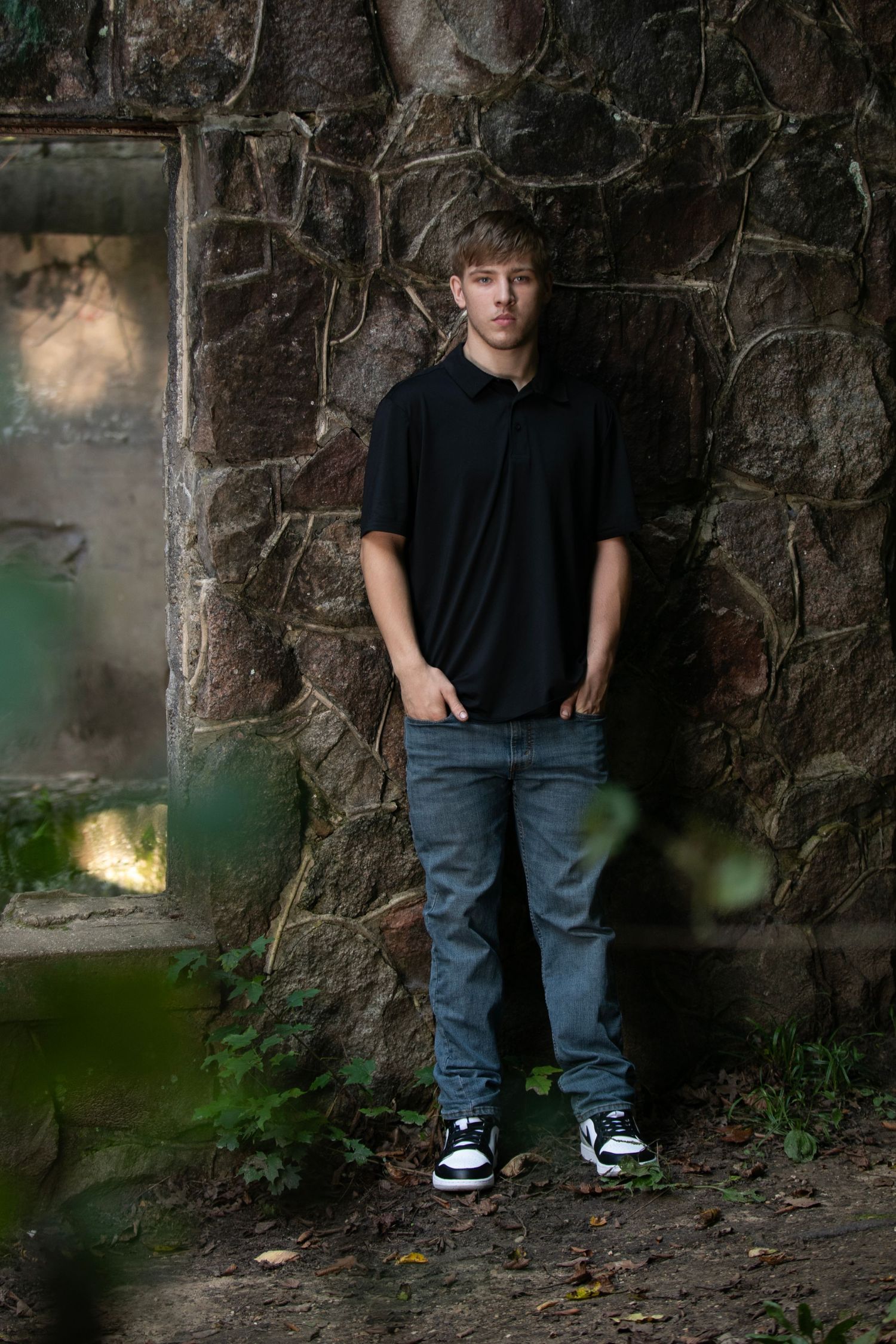 A person in casual black shirt and jeans stands against a rustic stone wall with green foliage in the foreground.