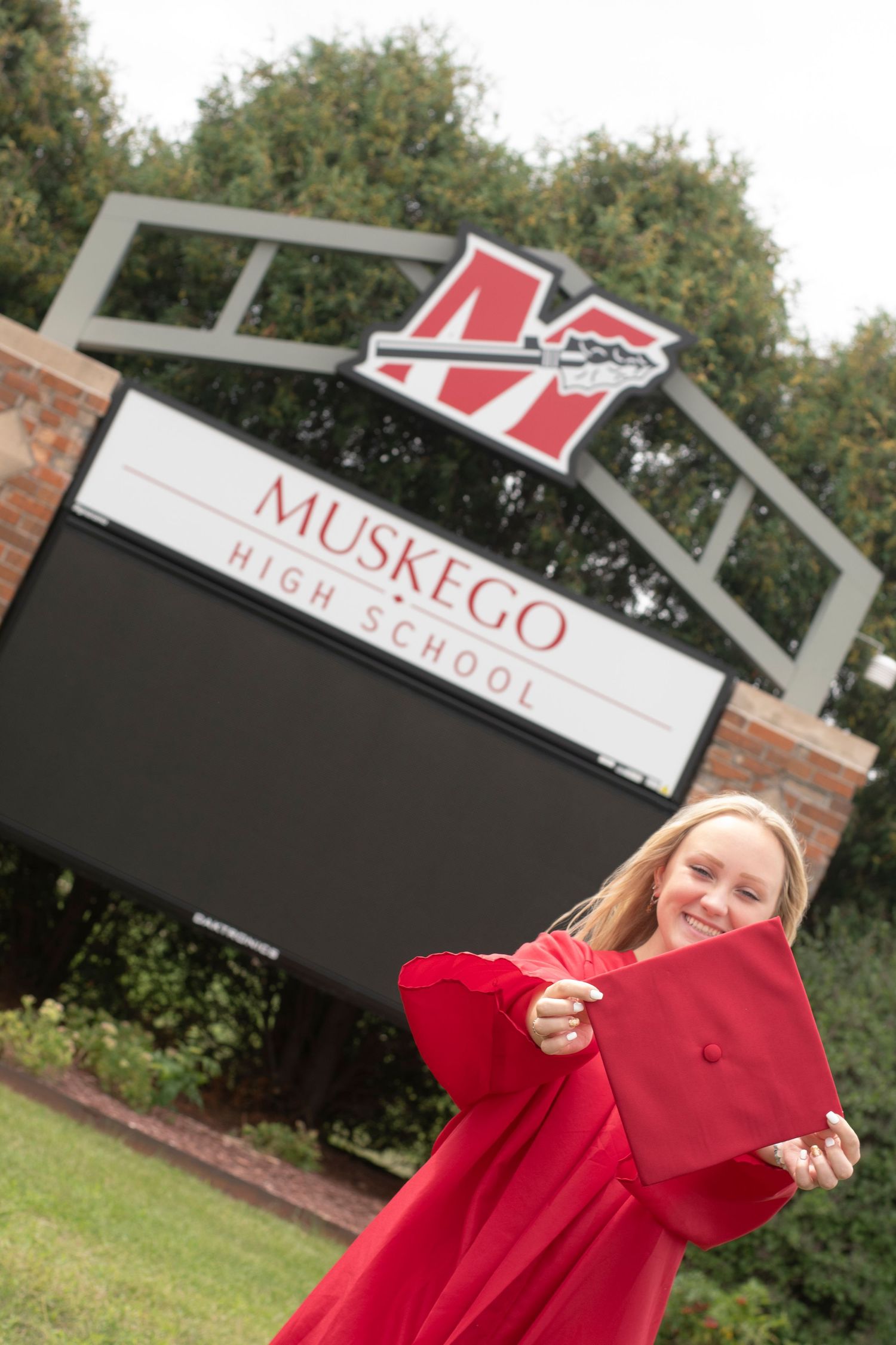 Figure holds red graduation cap outdoors with school signage visible in background.
