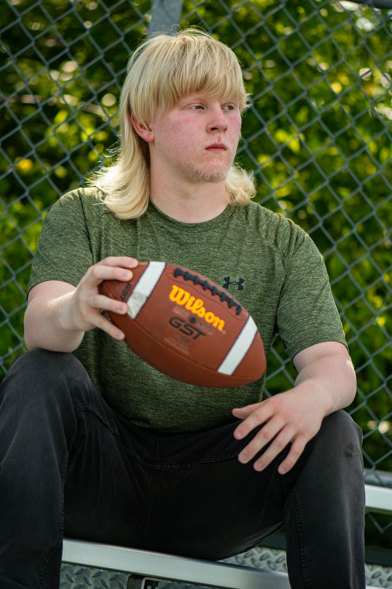 Person in olive green shirt sitting outdoors holding Wilson football against chain link fence. Game Day to Grad Day Seniors