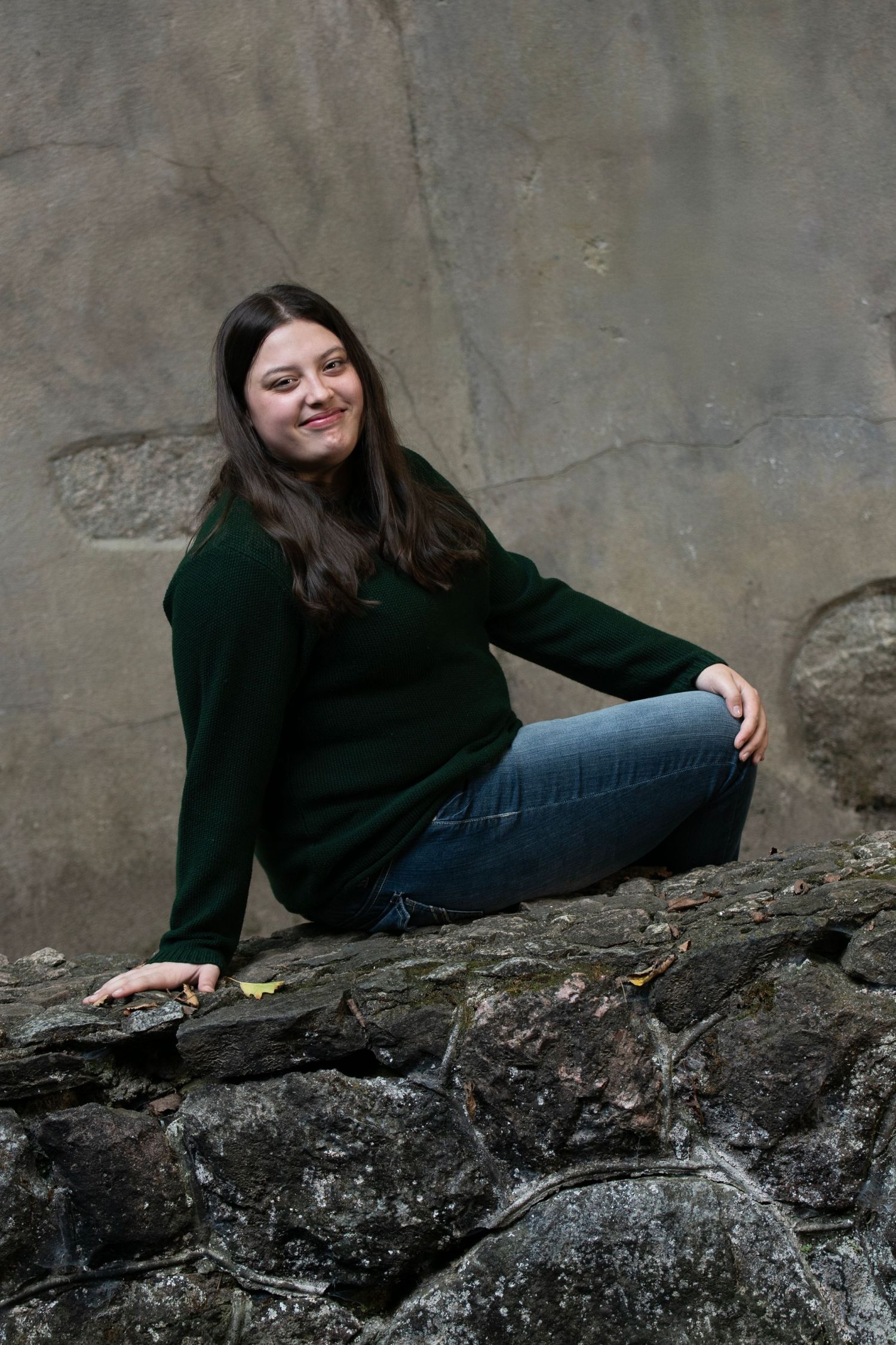 A person in a black sweater poses casually on a stone wall against a textured gray background.