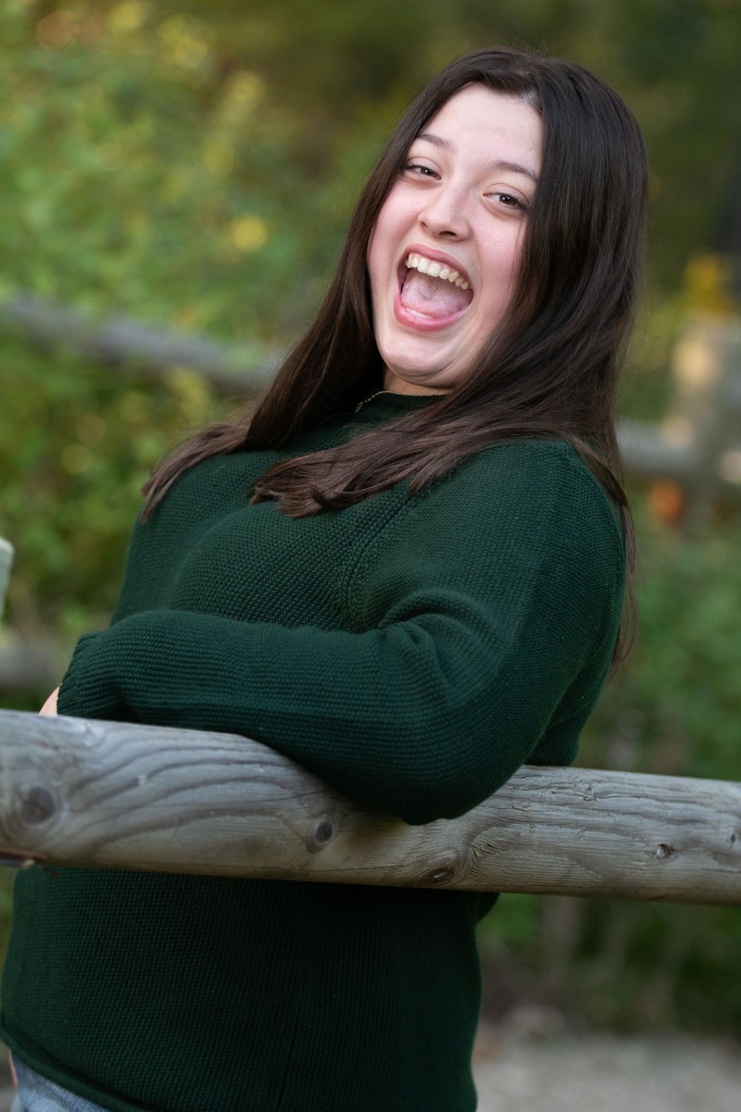 A person in a dark green sweater laughs while leaning back on a wooden log outdoors.