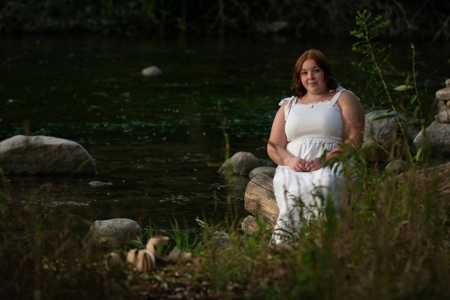 Woman in white dress sitting by a rocky riverbank surrounded by lush green vegetation.