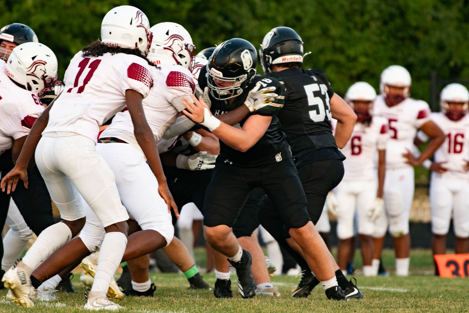 High school football players in black and white uniforms clash during an intense line scrimmage on a green field.