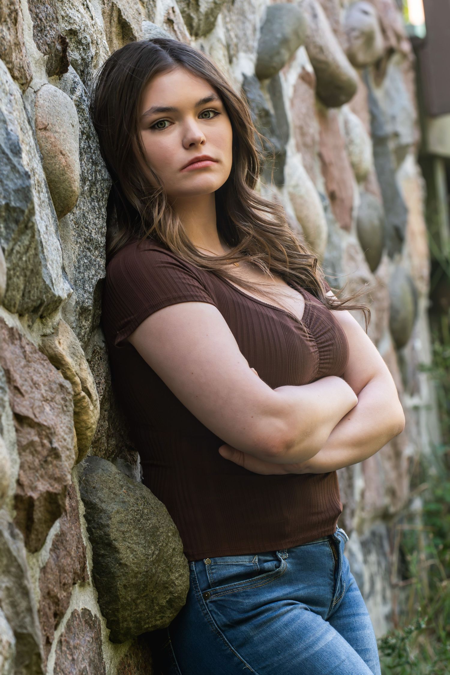 Young woman with brown hair in dark top and jeans leans against rustic stone wall with arms crossed.