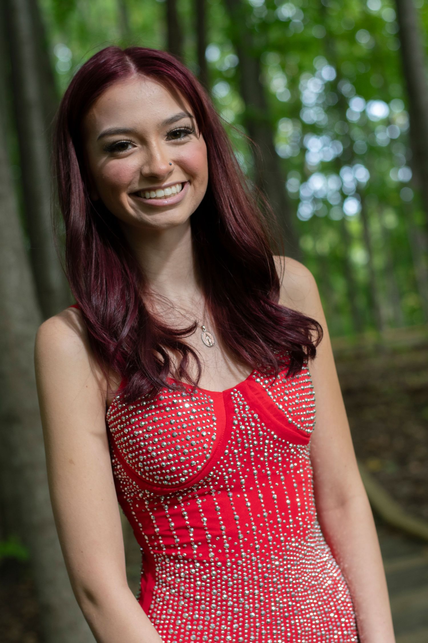 Smiling young woman with red hair wearing a sparkly red dress, posing outdoors in a lush green forest.