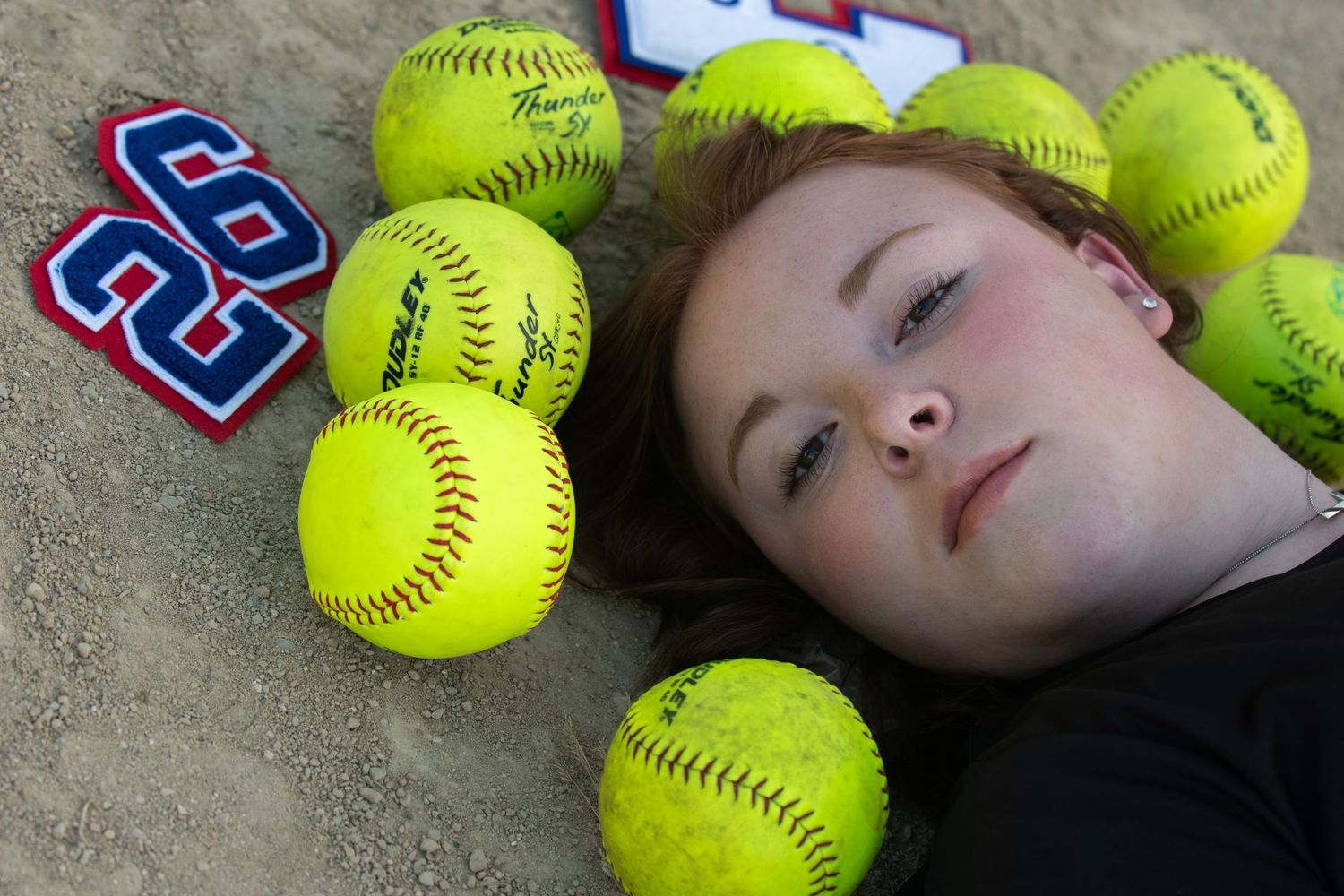 Softballs are arranged on a dirt surface next to a senior lying down for a creative senior portrait experience with Simply Junique portraits
