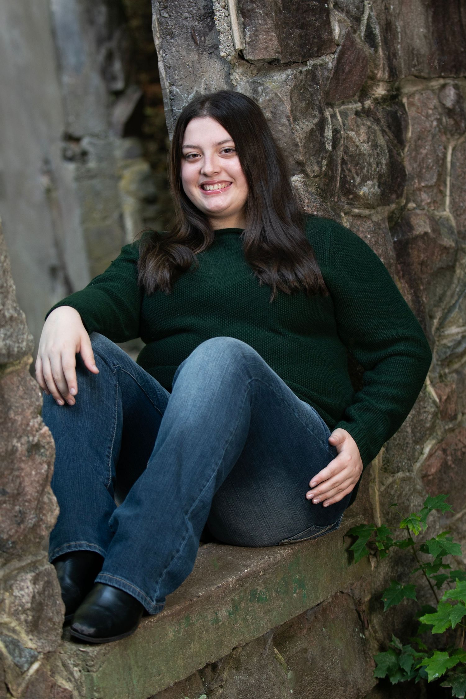 A person in a dark green sweater and jeans sits casually on a stone wall with ivy growing nearby.