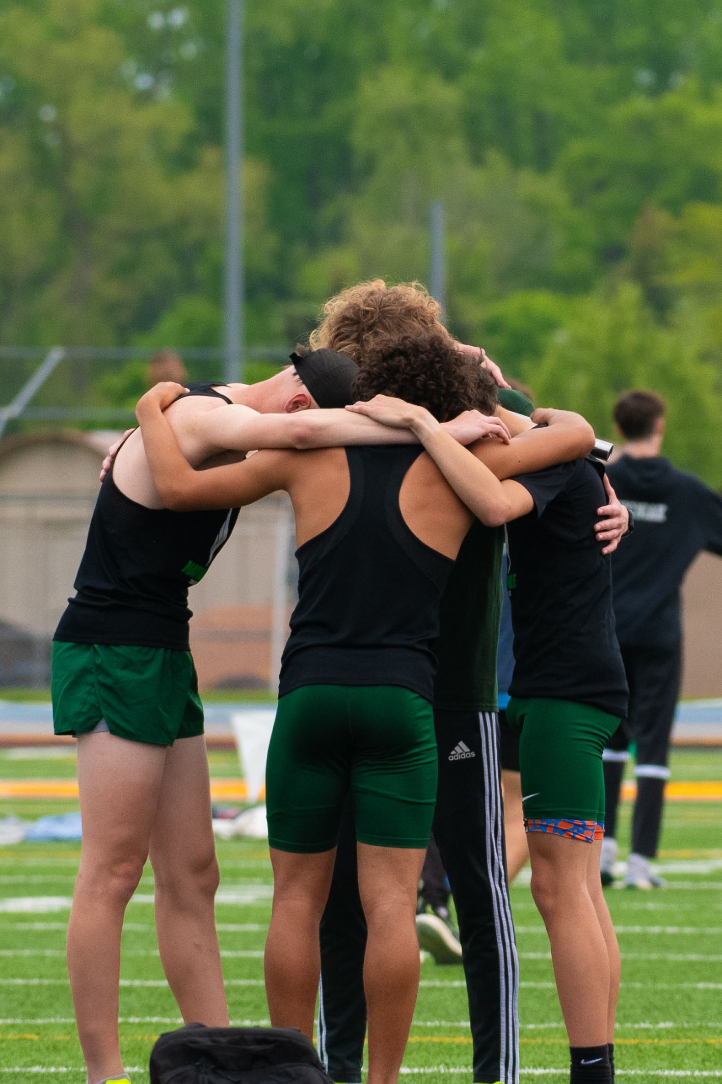 Track team members in green uniforms huddle together during practice on an outdoor field.