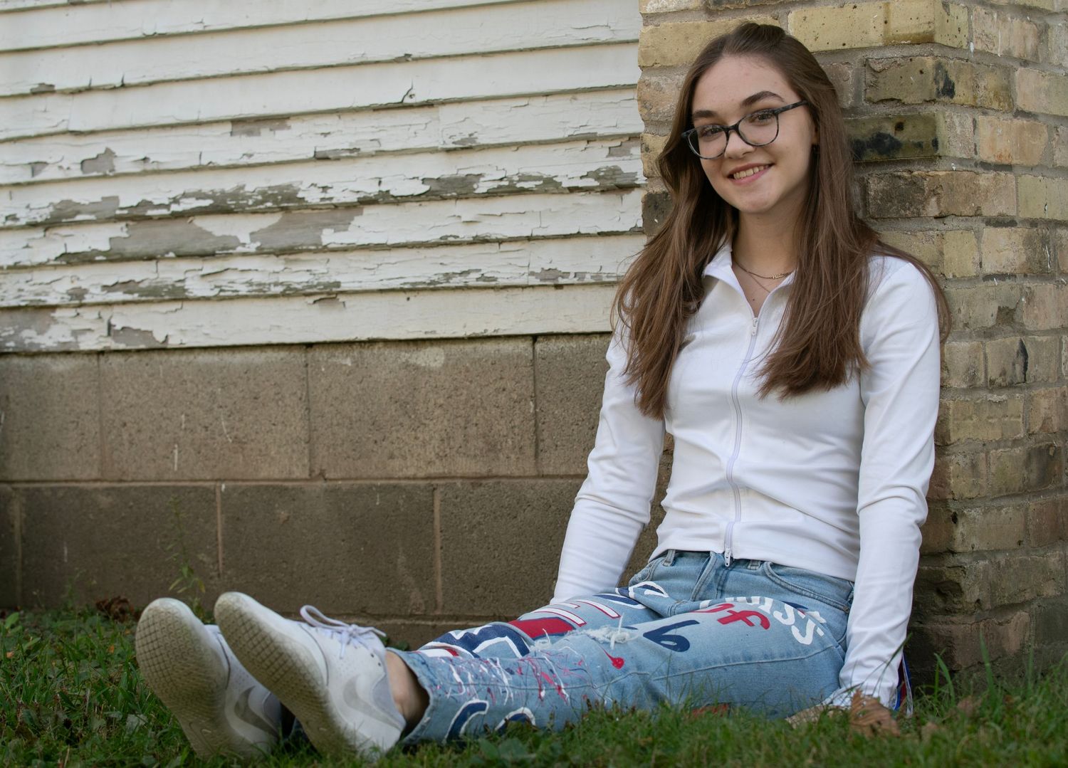 A person in a white shirt and jeans sits casually on grass near a weathered brick wall.