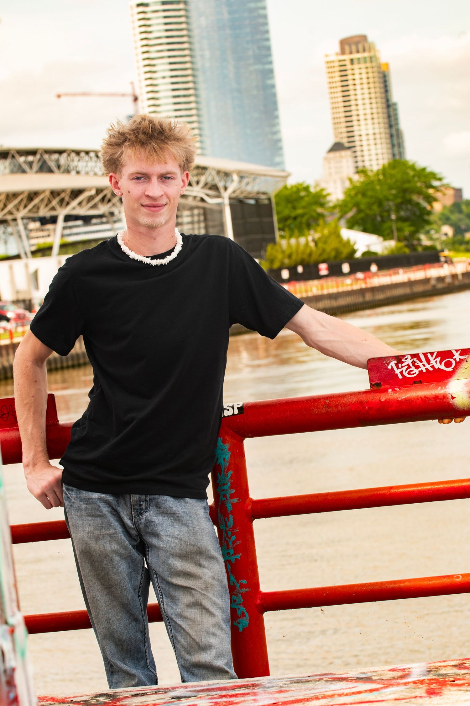 Young man in black t-shirt leaning on red railing by urban waterfront with city skyline background.