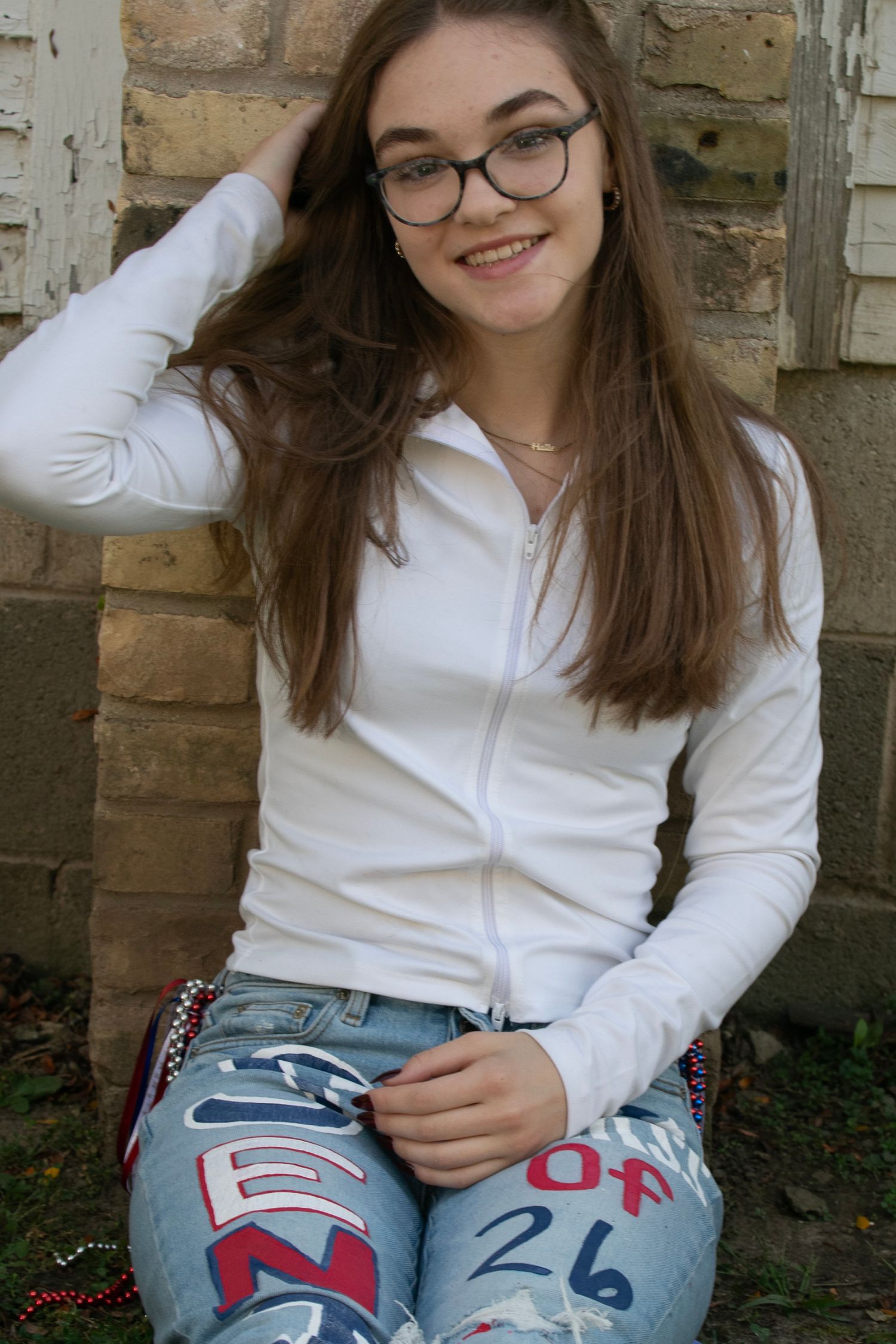 A smiling teenage girl with glasses wearing a white zip-up hoodie and painted jeans sits against a brick wall.