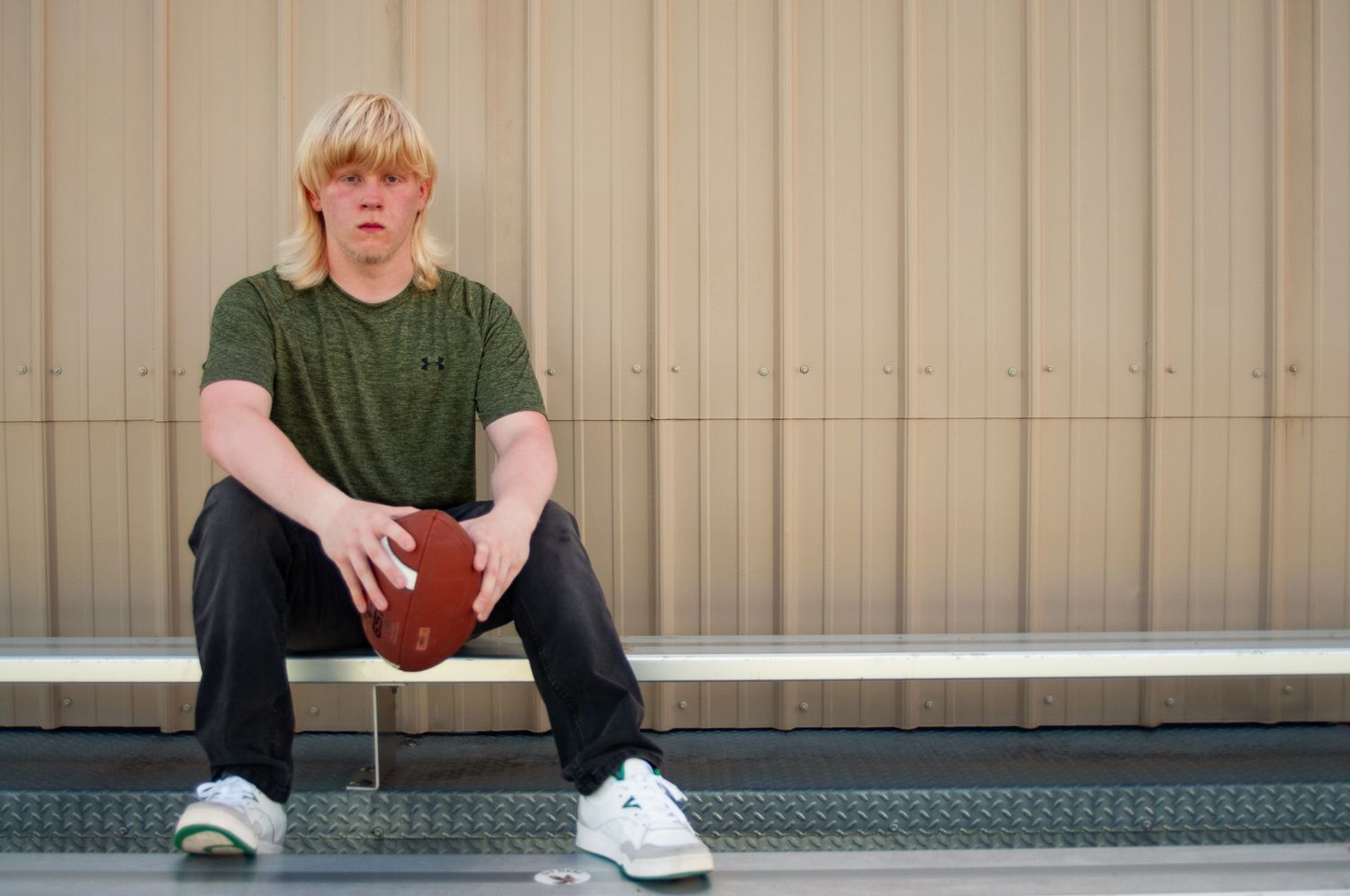 Person in dark clothing sitting on metal bleachers holding football. Game Day to Grad Day Senior Portrait Experience with Simply Junique