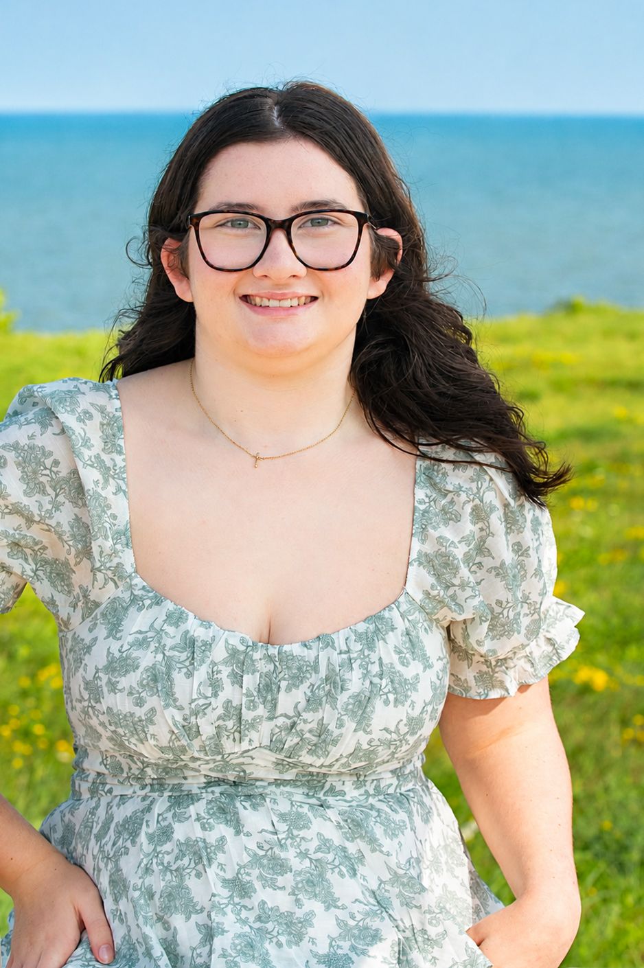 Young woman with dark hair and glasses smiling outdoors in a floral dress with ocean and green field behind her.