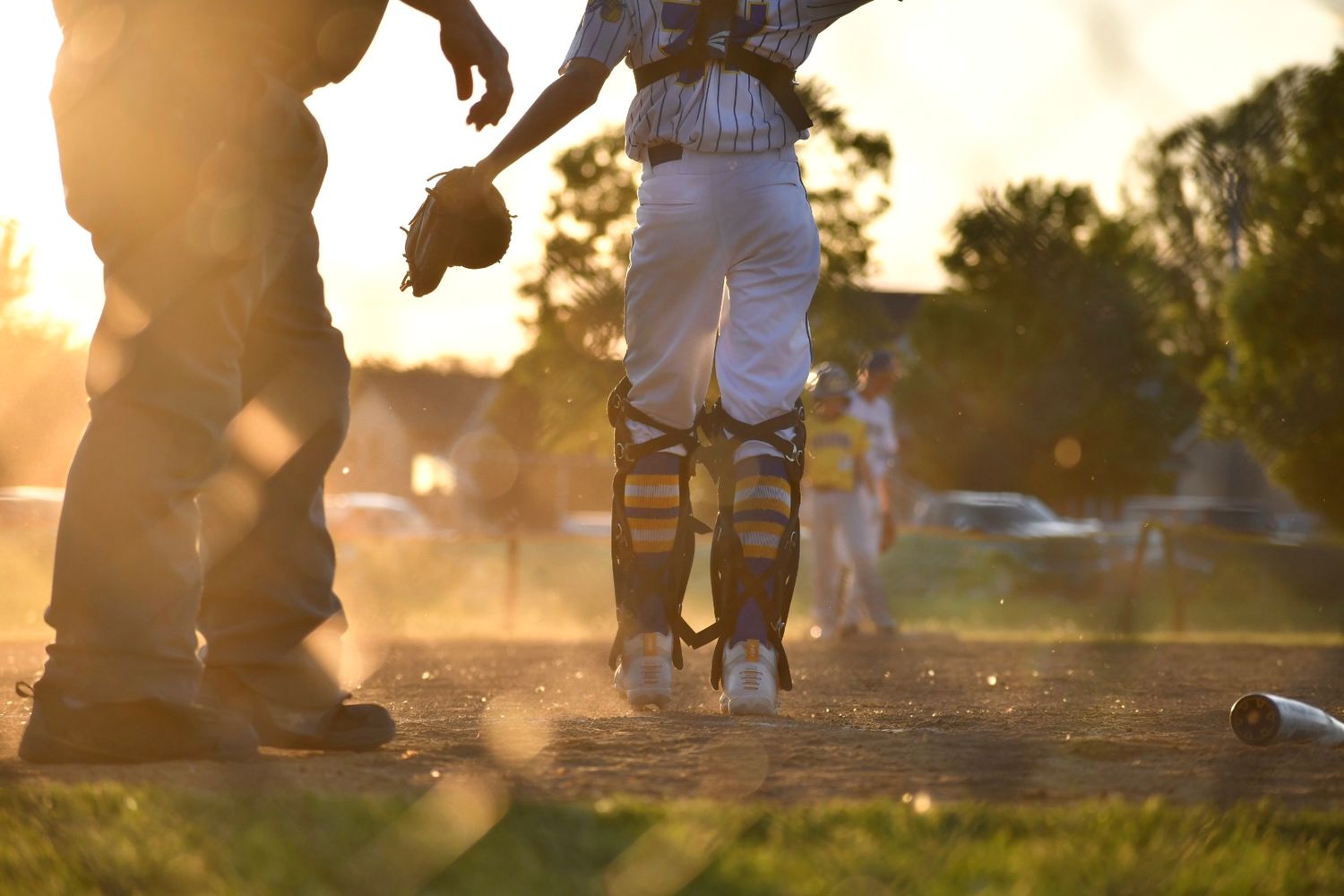 Baseball catcher in full gear stands on dusty field at golden sunset during a youth baseball game.