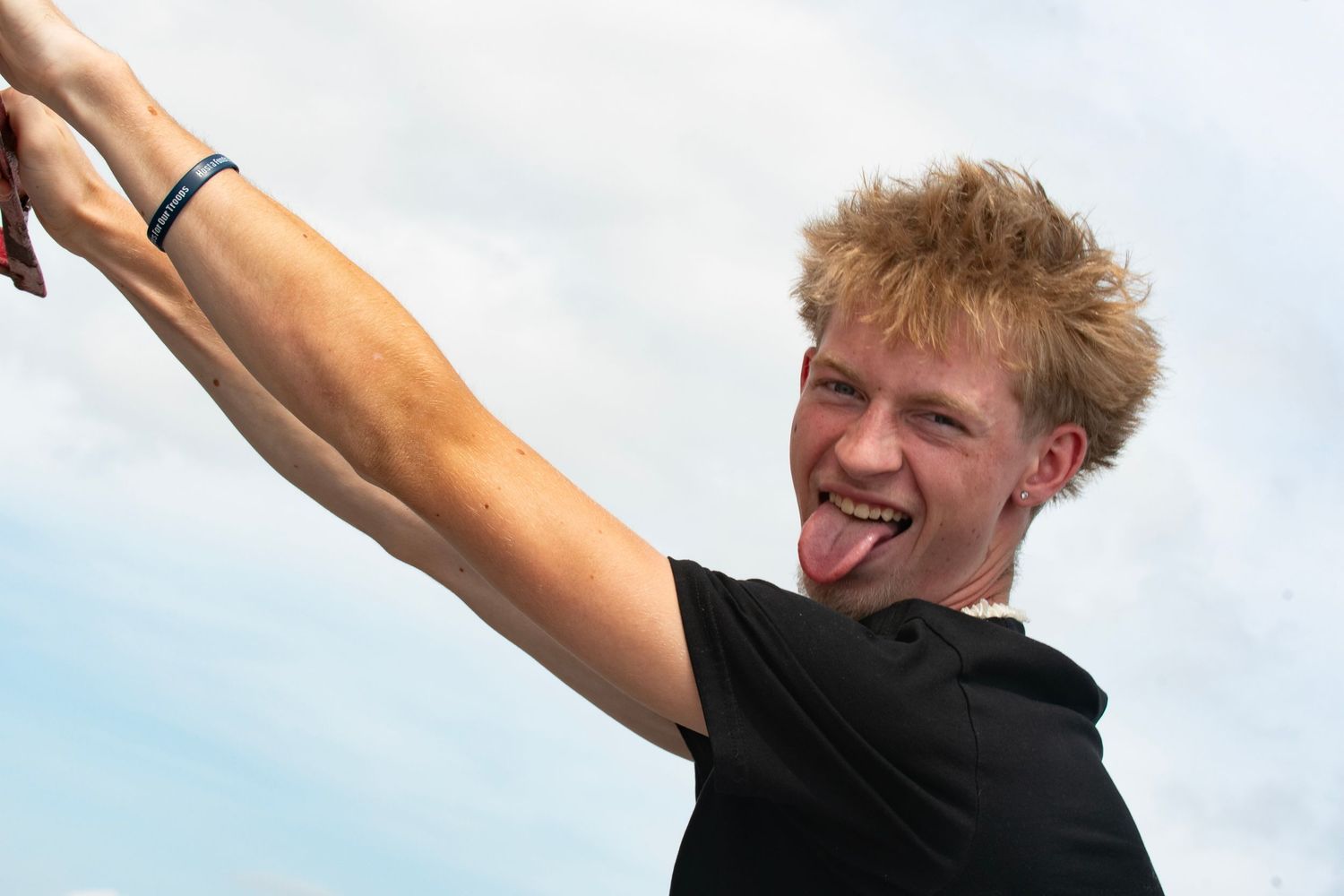 Young man with blonde hair sticking out tongue while taking a selfie outdoors against a cloudy sky.