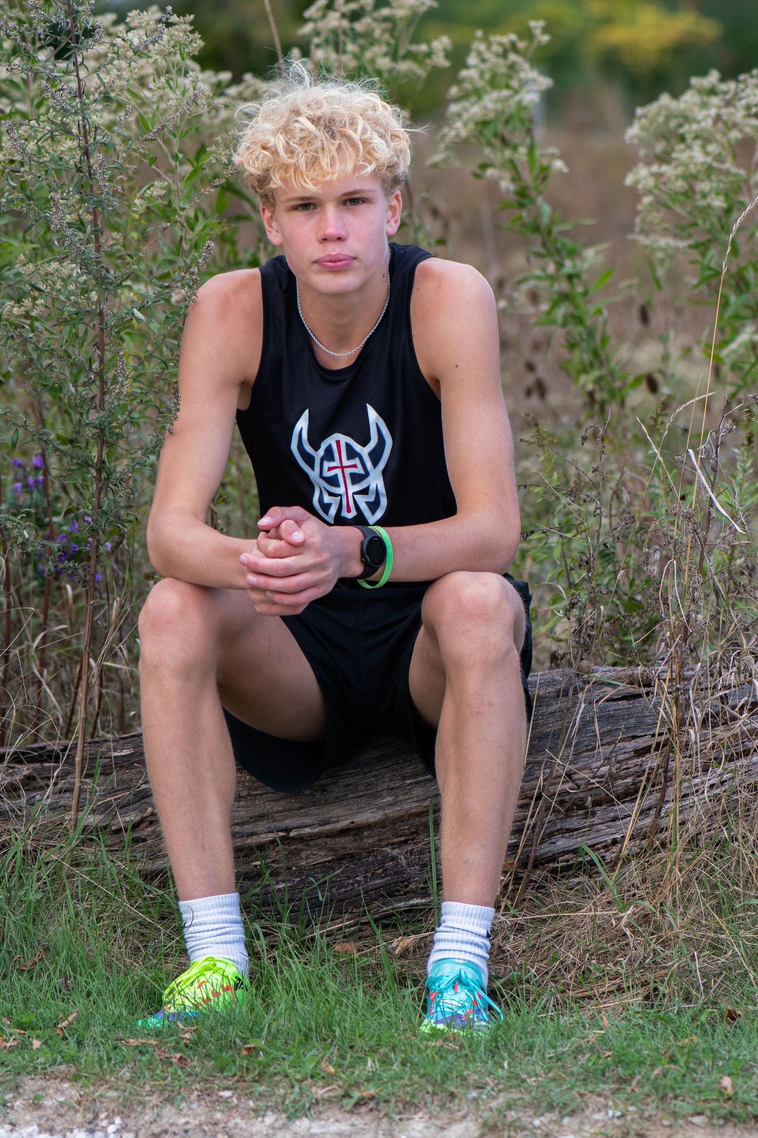 A person sits on a fallen log in a wooded area wearing a black tank top with a Viking logo and colorful shoes. Milwaukee County Grounds