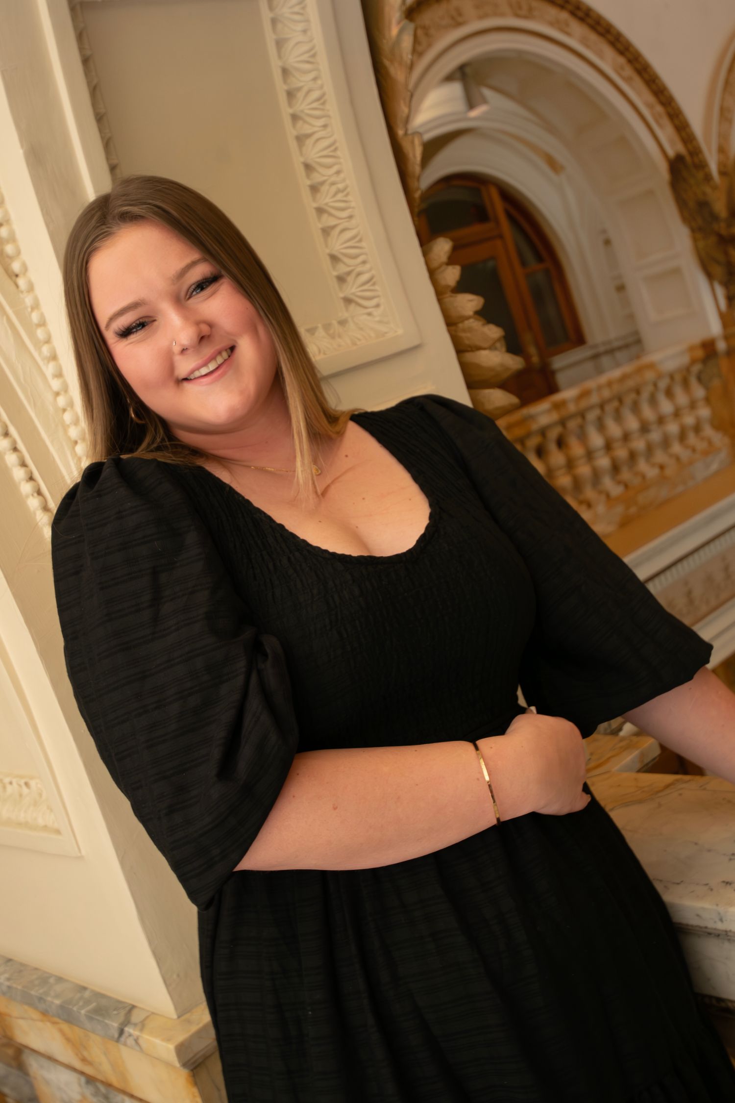 Person in a black dress poses against an ornate white pillar in an elegant interior setting.