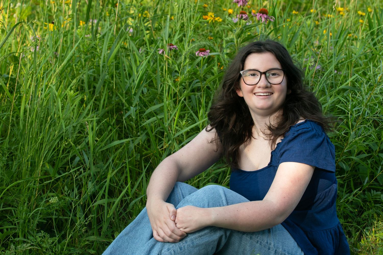 Young woman with glasses sitting in a lush green meadow with wildflowers, smiling outdoors.