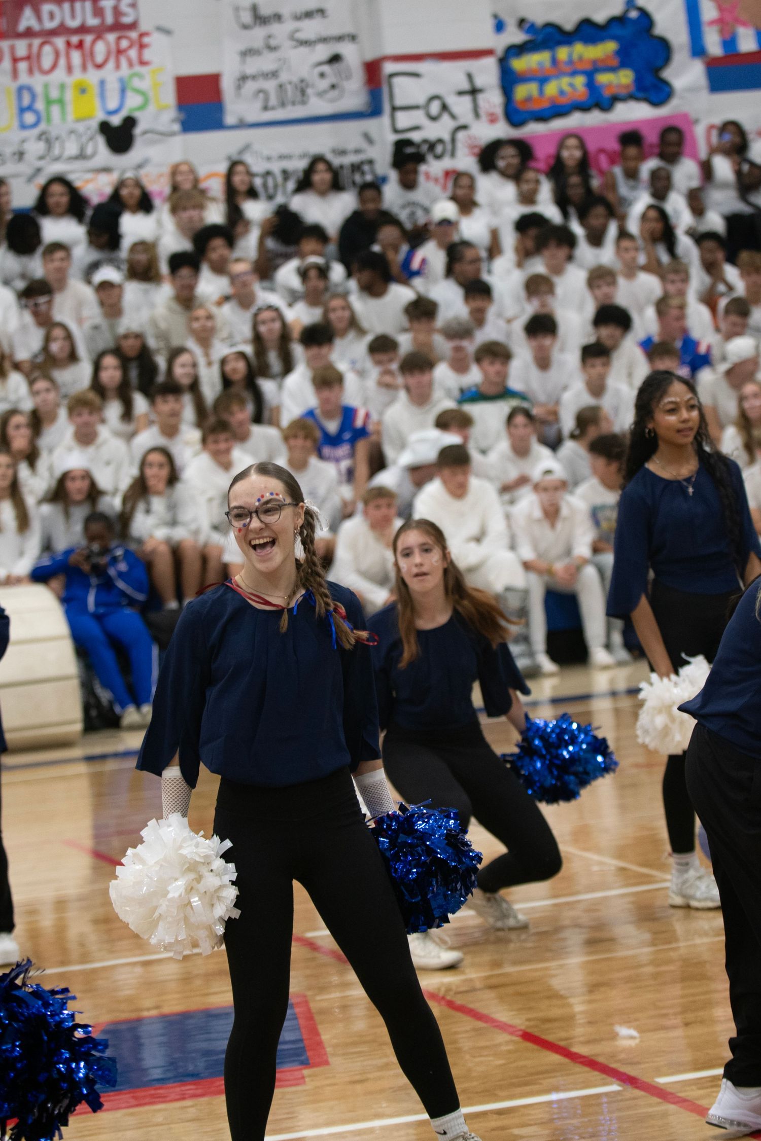 A cheerleader in navy blue performs with pom-poms at a school pep rally with students in white filling the bleachers.