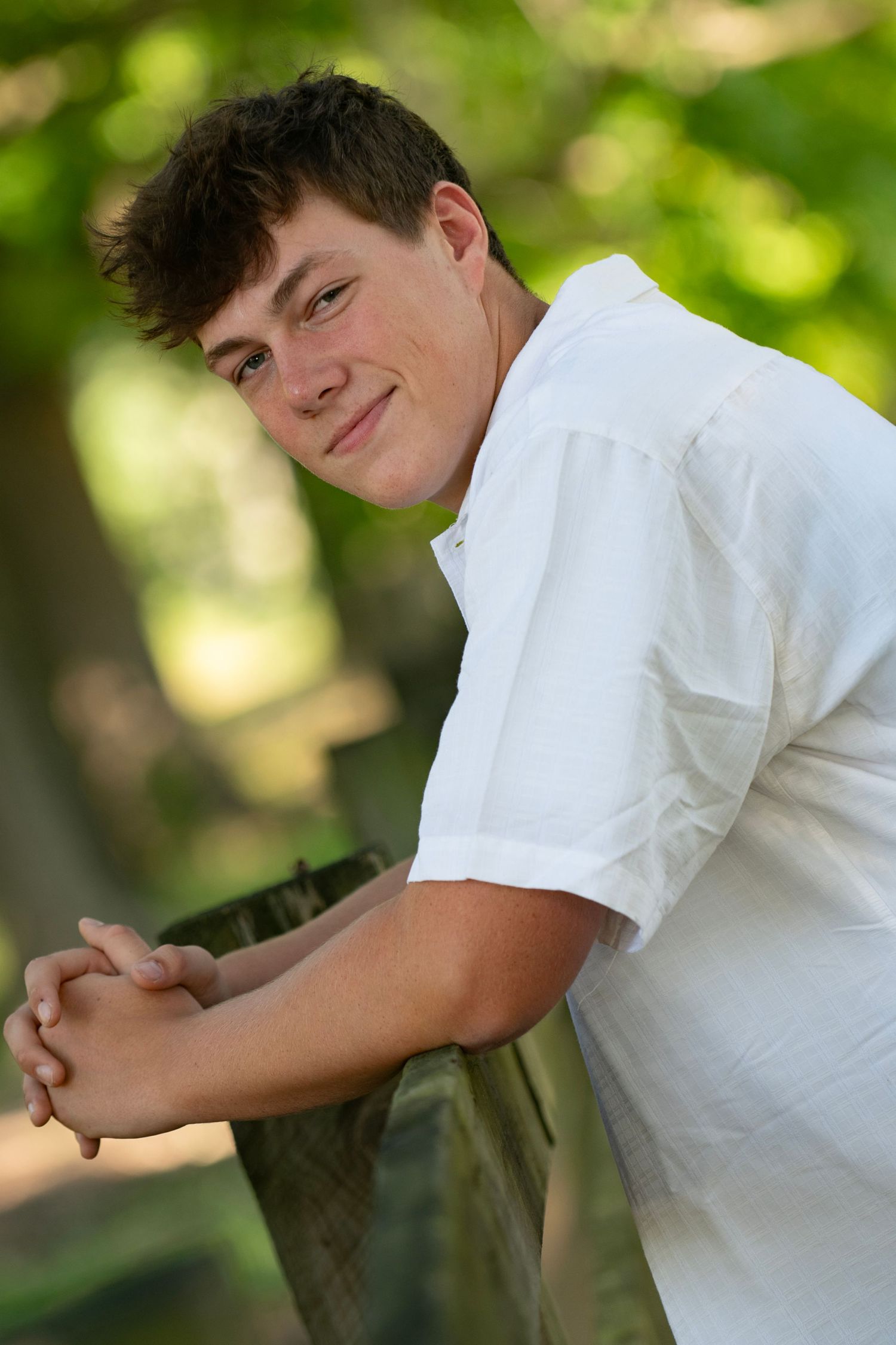 Young man in white shirt leaning on fence, smiling outdoors with green trees in background.