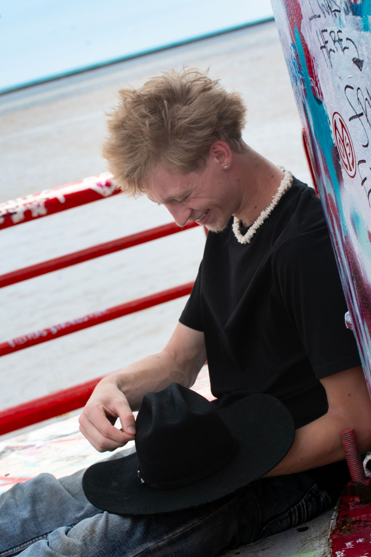 Young blond man in black t-shirt holding a black hat, sitting outdoors near red railings with graffiti.