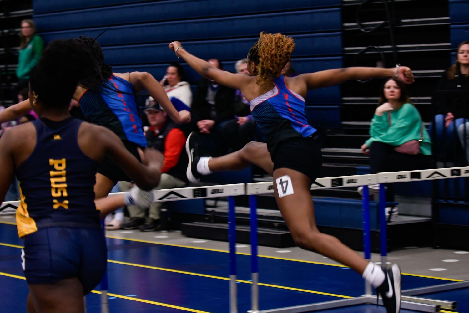 Track athletes clearing hurdles during an indoor competition.