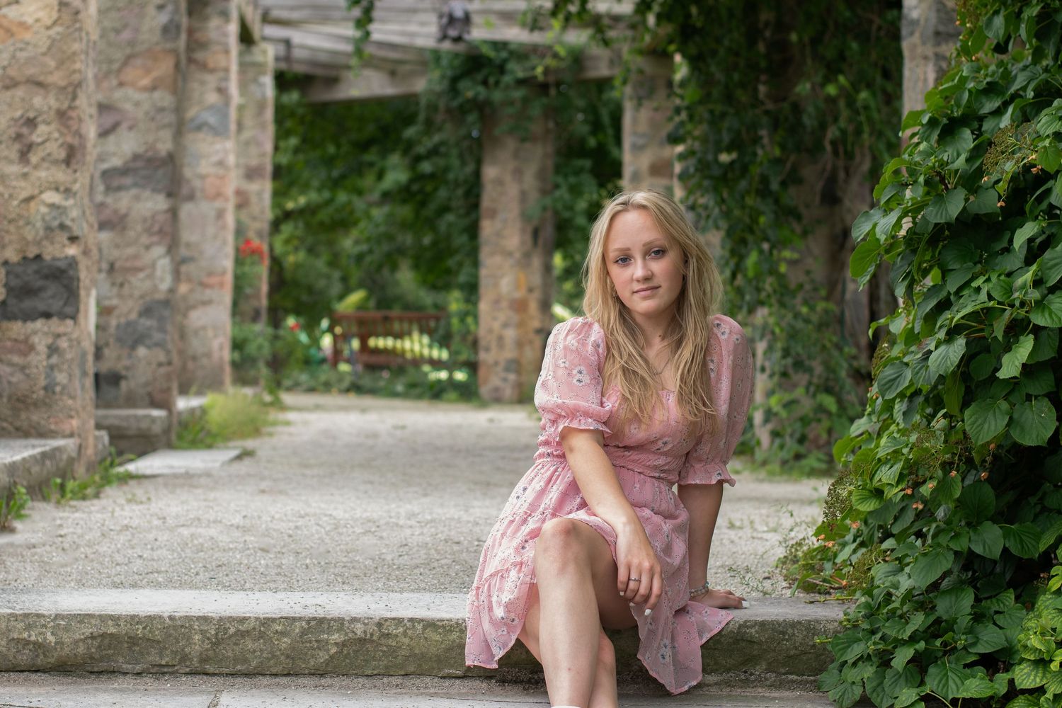 Outdoor portrait series in pink dress sitting on stone steps surrounded by greenery.