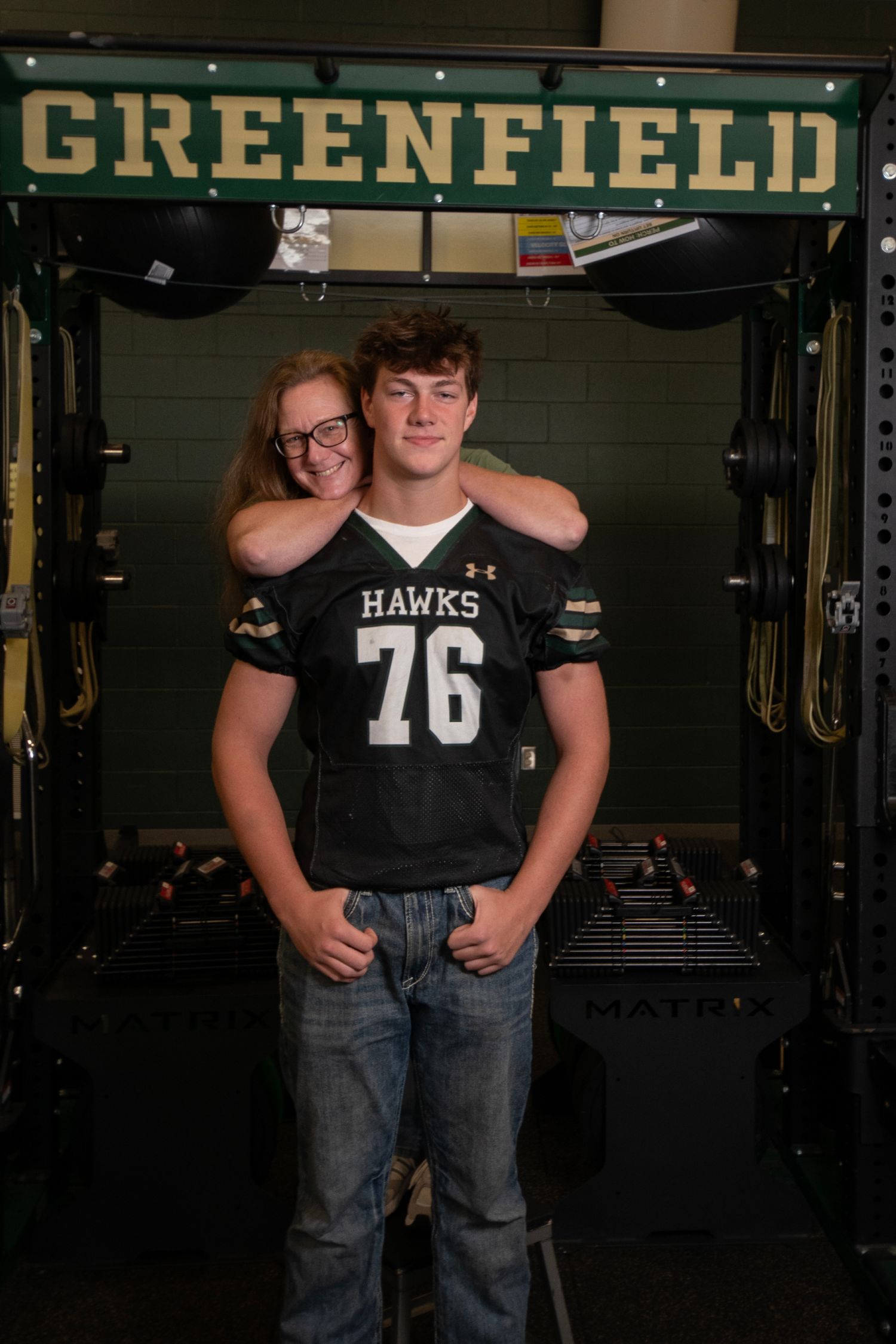 Student athlete wearing Hawks jersey and jeans poses in front of Greenfield athletic facility sign.