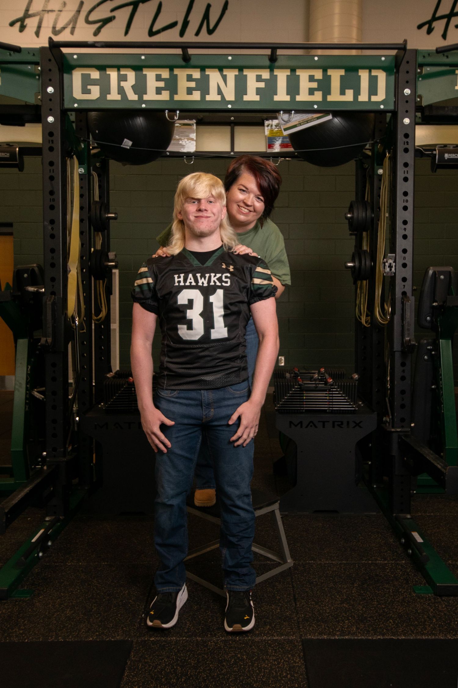 Two people stand together in a gym weight room with 'Greenfield' text visible on the wall behind them.