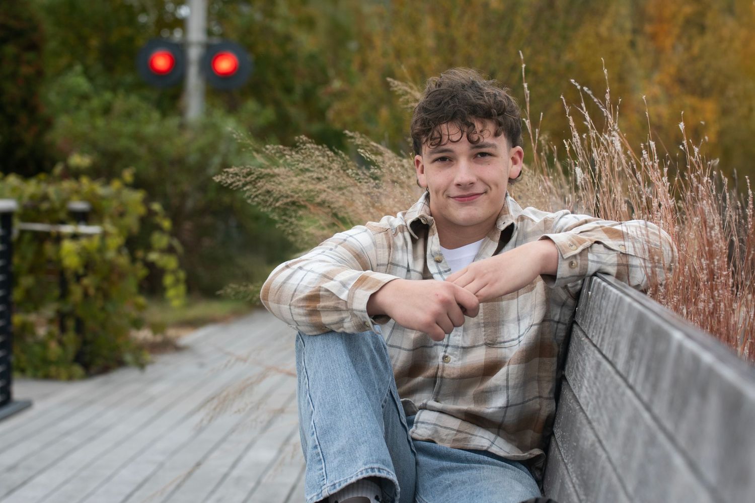 A teenage boy in a plaid shirt sits relaxed on a wooden bench outdoors, surrounded by autumn foliage.