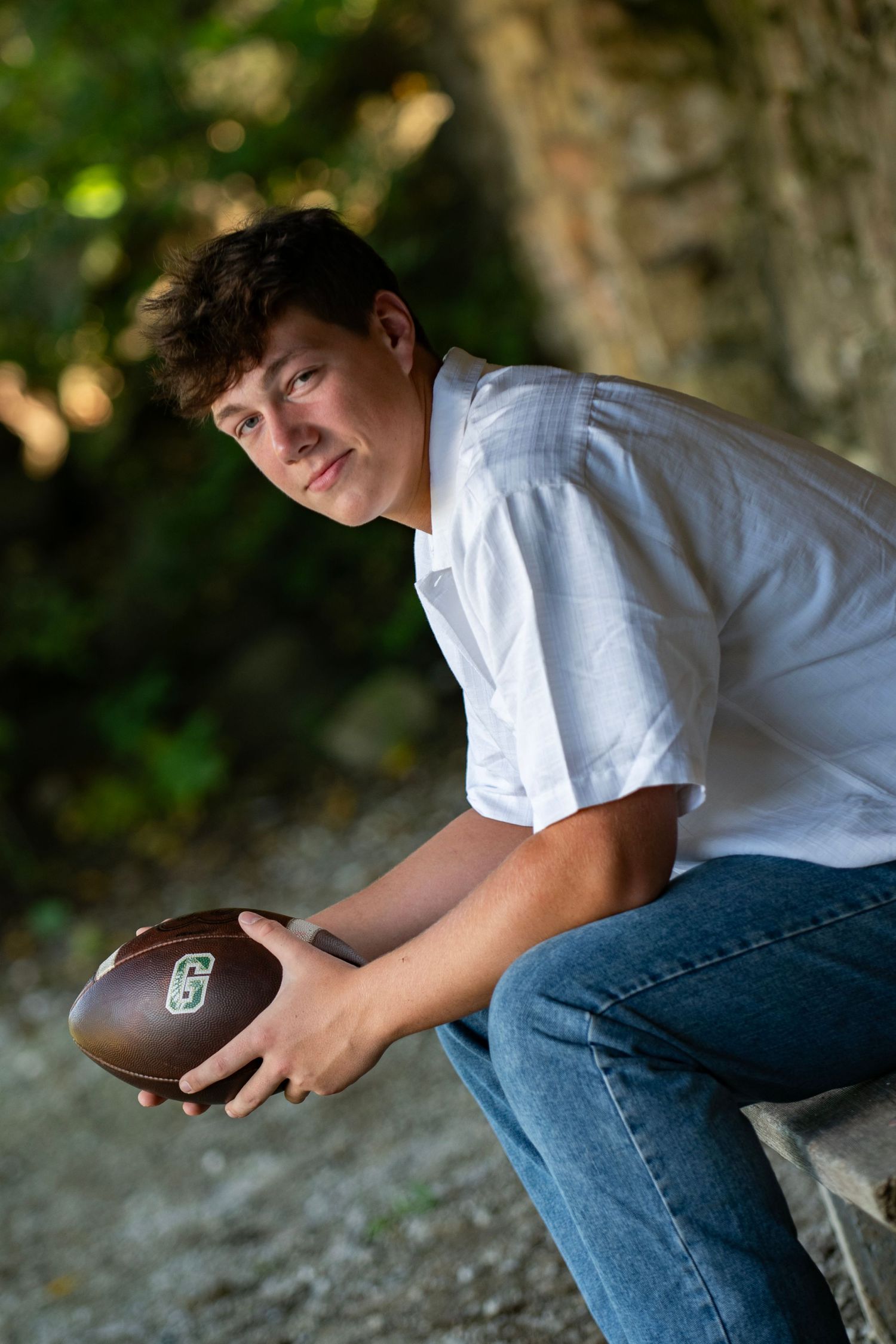 Teen boy in white shirt holding football while sitting outdoors, smiling at camera during senior portrait session.